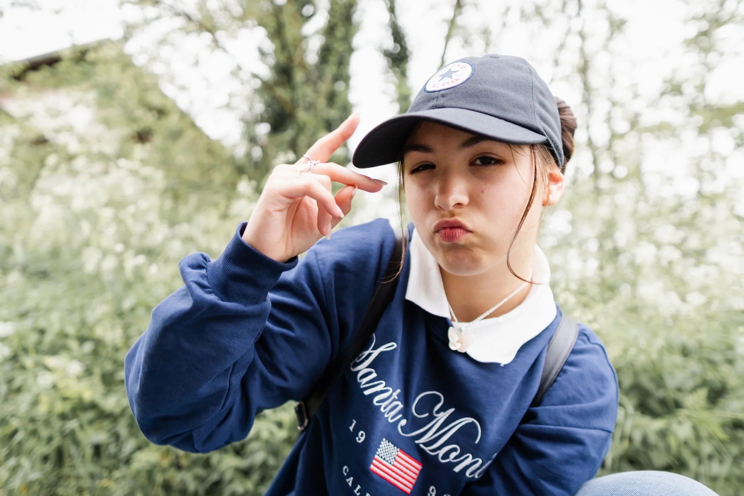 Young girl outdoors making a playful face, wearing a baseball cap, navy sweatshirt with American flag and text, and a backpack.