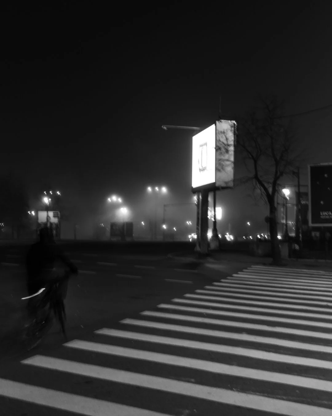 A black and white nighttime scene of a street with a crosswalk in the foreground and an illuminated billboard in the background. A person is visible crossing the street on the left side.