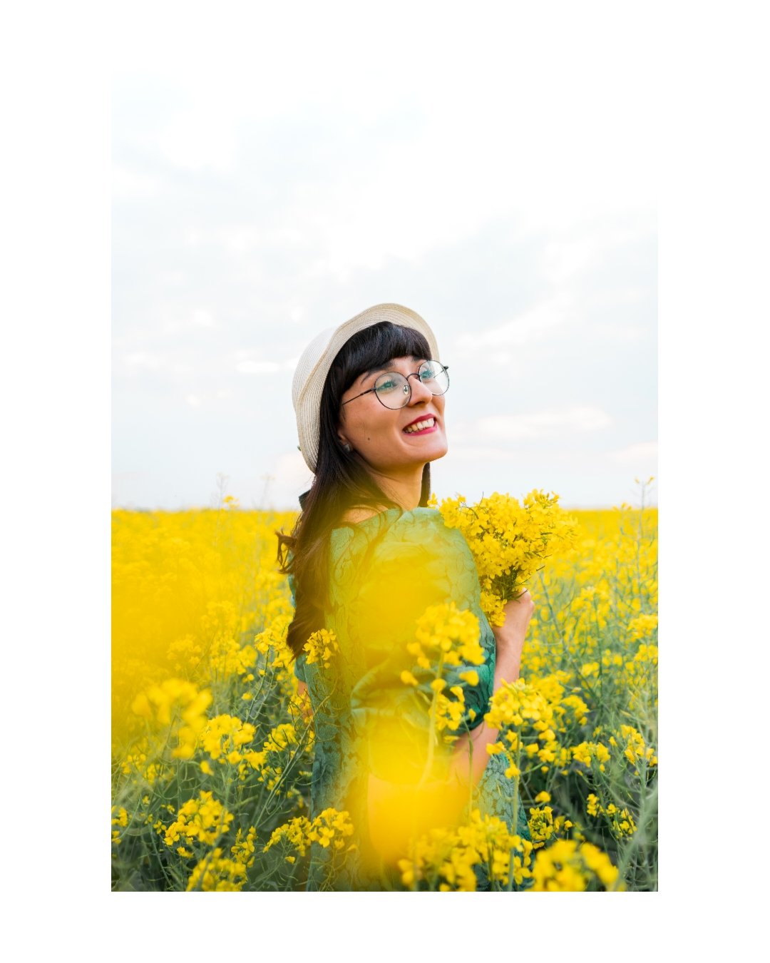 A woman wearing glasses, a hat, and a blue dress stands in a yellow flower field, smiling and holding a bouquet of yellow flowers.