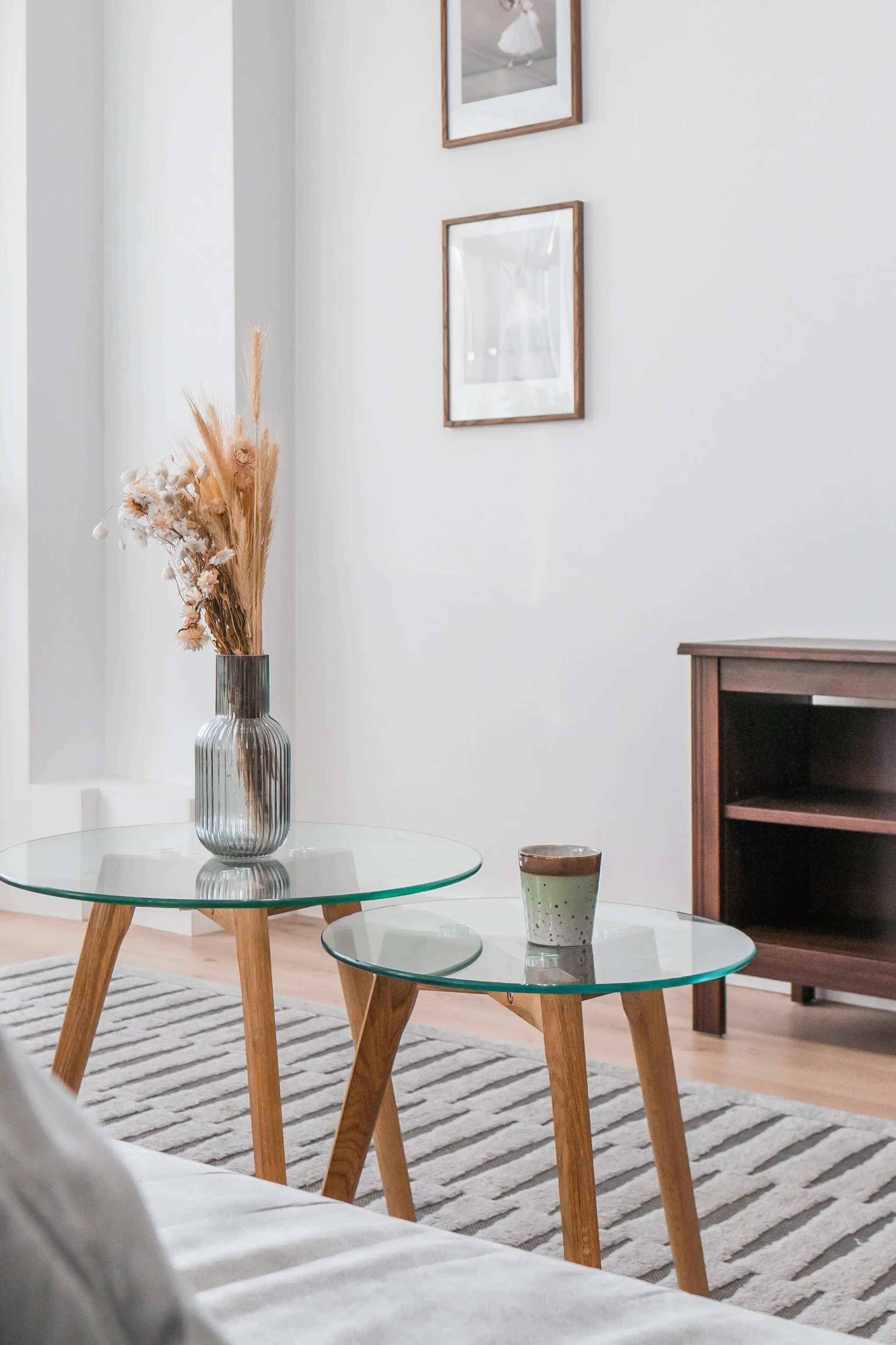 Living room with two overlapping glass-top tables with wooden legs, a vase with dried flowers, a small cup, a striped rug, wall art, and a wooden console against a white wall.