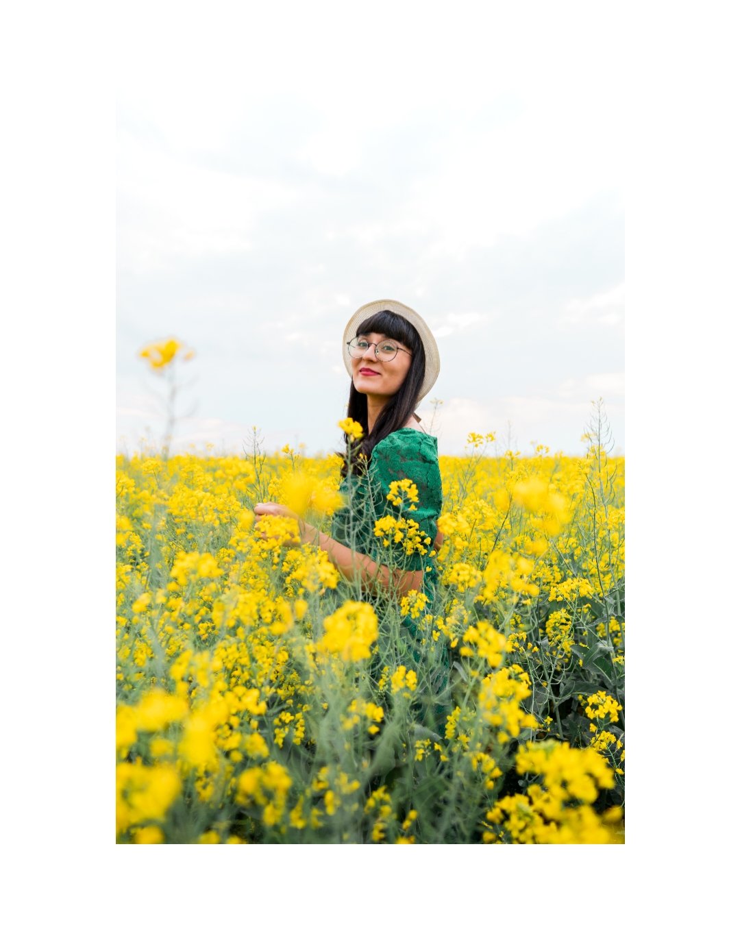 A woman wearing glasses and a straw hat stands in a yellow flower field under a cloudy sky.