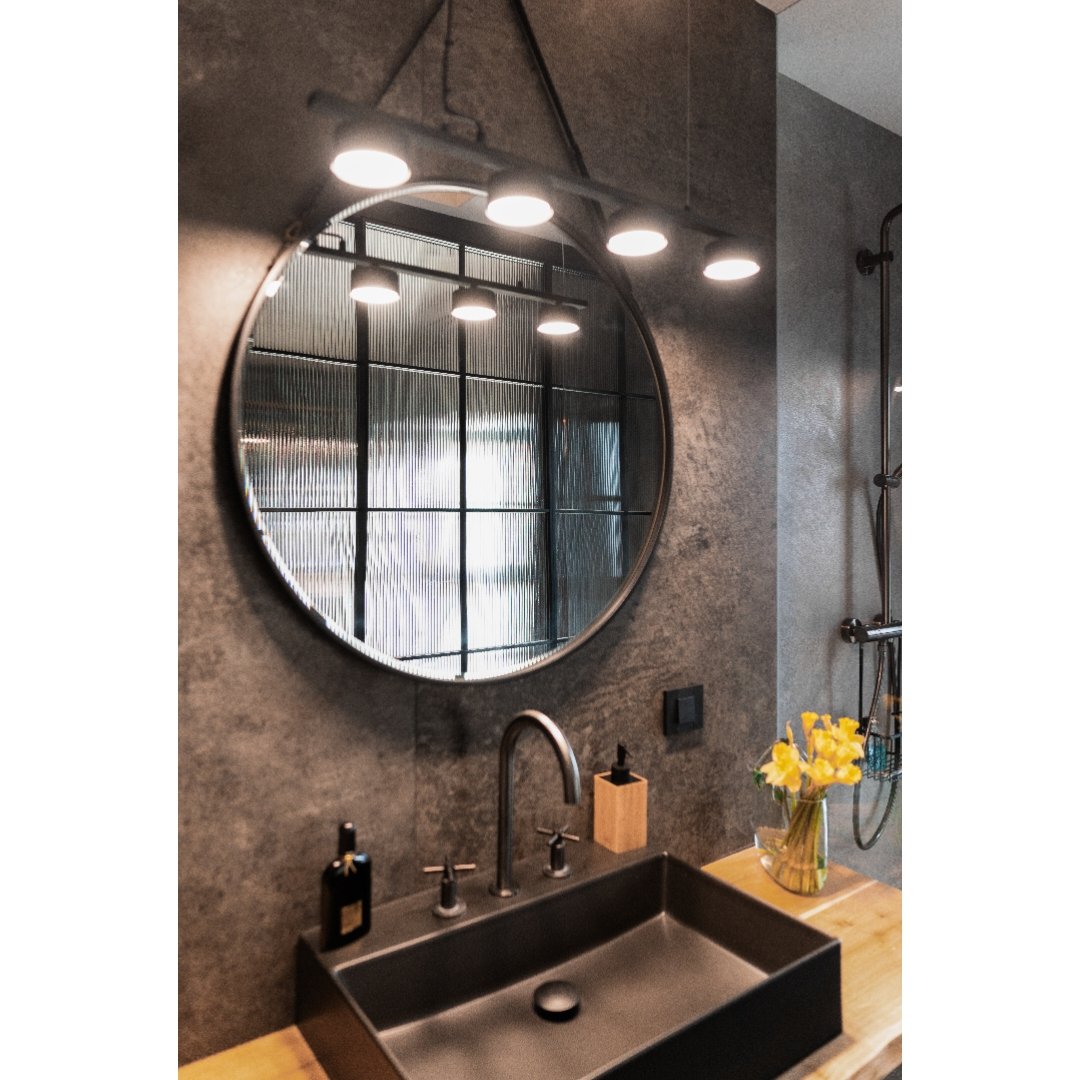 Bathroom with a round mirror, black sink with modern faucet, a small black soap dispenser, a wooden box for toiletries, and a glass vase with yellow flowers on a wooden countertop. The wall is textured gray, and a shower with a glass door, showerhead, and gray tiles is visible in the background.