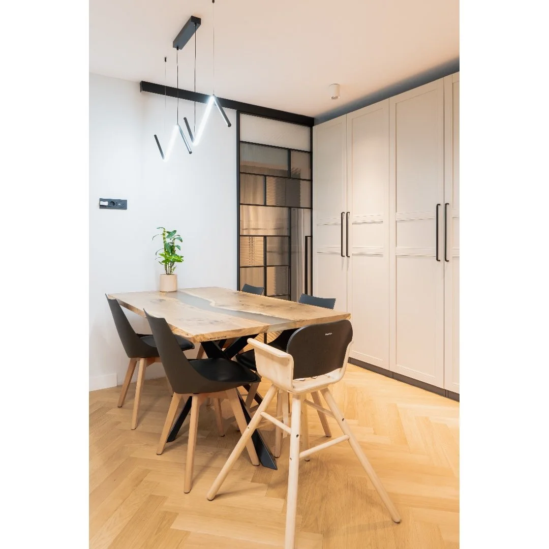 Modern dining room with a wooden table, black and beige chairs, a small plant, built-in white cabinets, and a black wall with glass storage panels, illuminated by stylish hanging lights.
