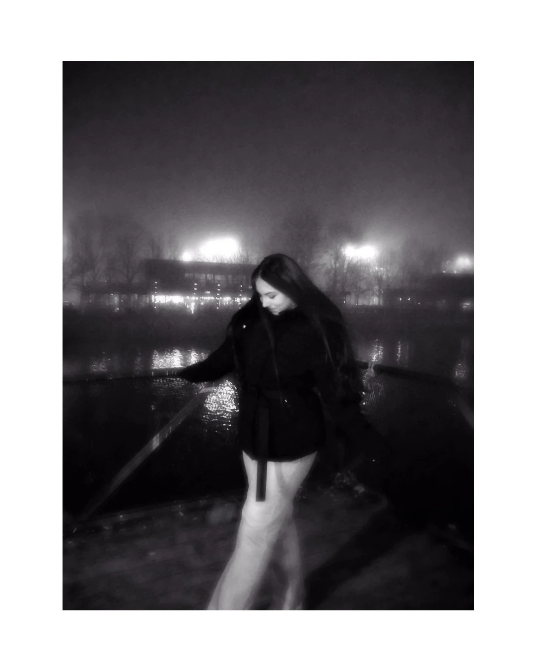 A black-and-white photo of a young woman standing on a boat at night, wearing a black coat, with river and illuminated buildings in the background.