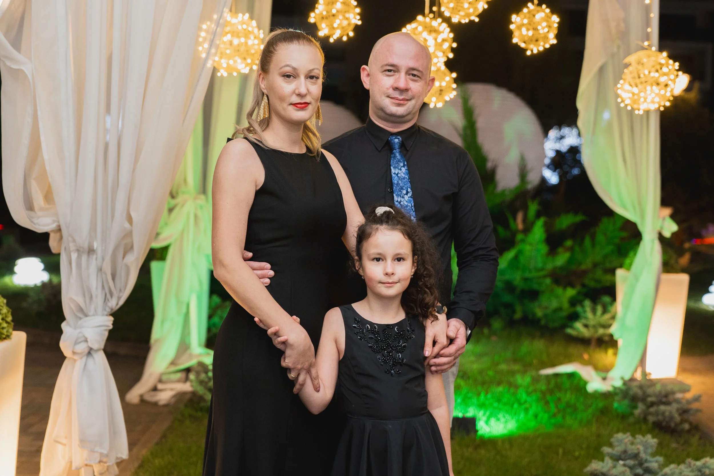 A family of three, a woman, a man, and a young girl, dressed in black and posing under decorative lights and curtains at an outdoor event at night.