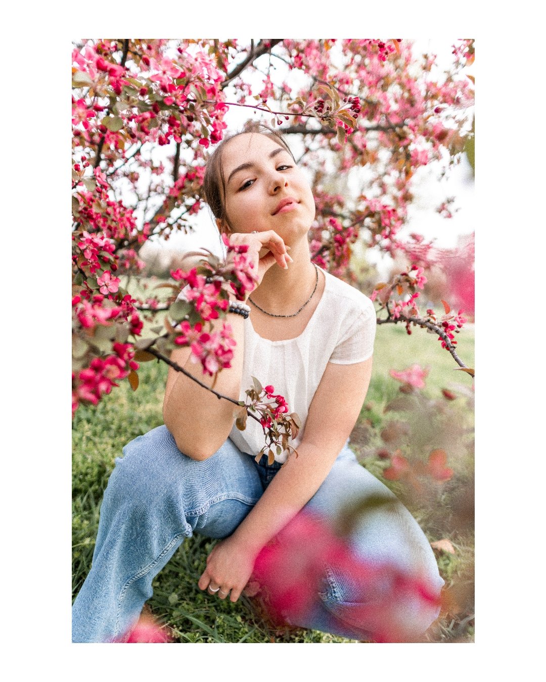 Young woman in white shirt and jeans sitting among pink flowering trees outdoors.