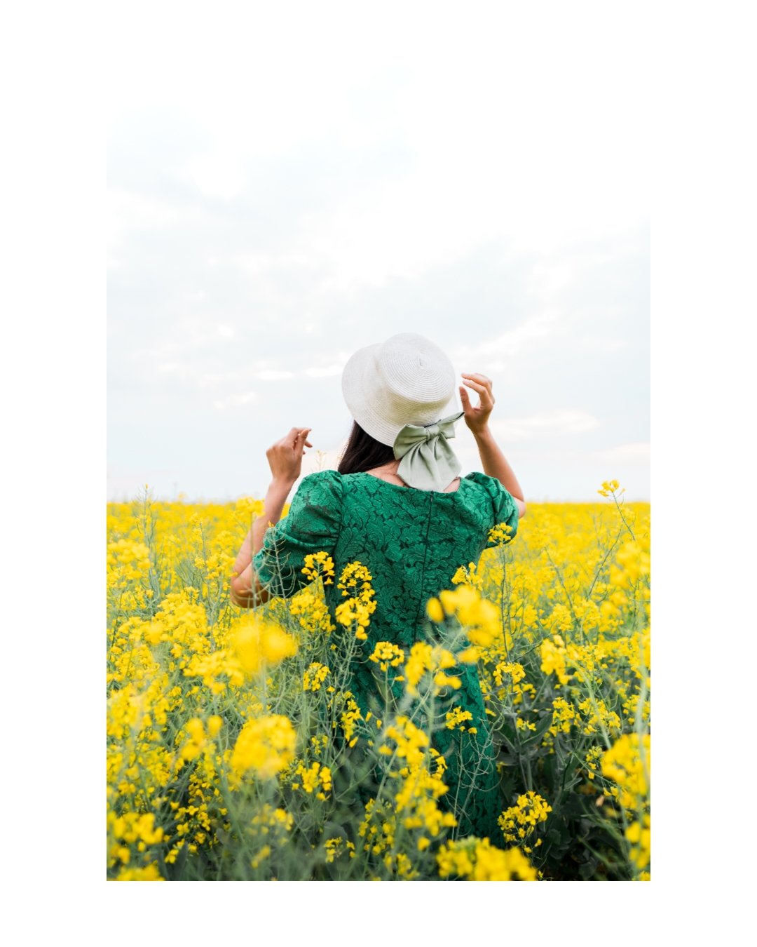 A woman in a green dress and white hat with a bow, standing in a yellow flower field under a cloudy sky.