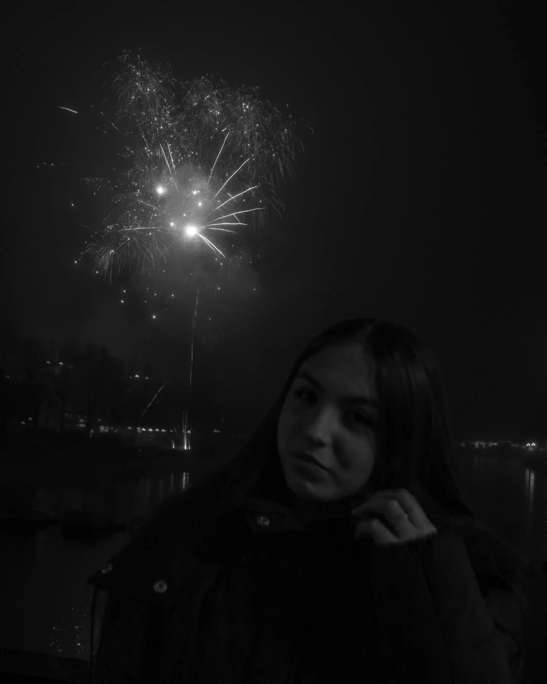 A woman posing in front of fireworks at night, with a dark sky and a body of water in the background.