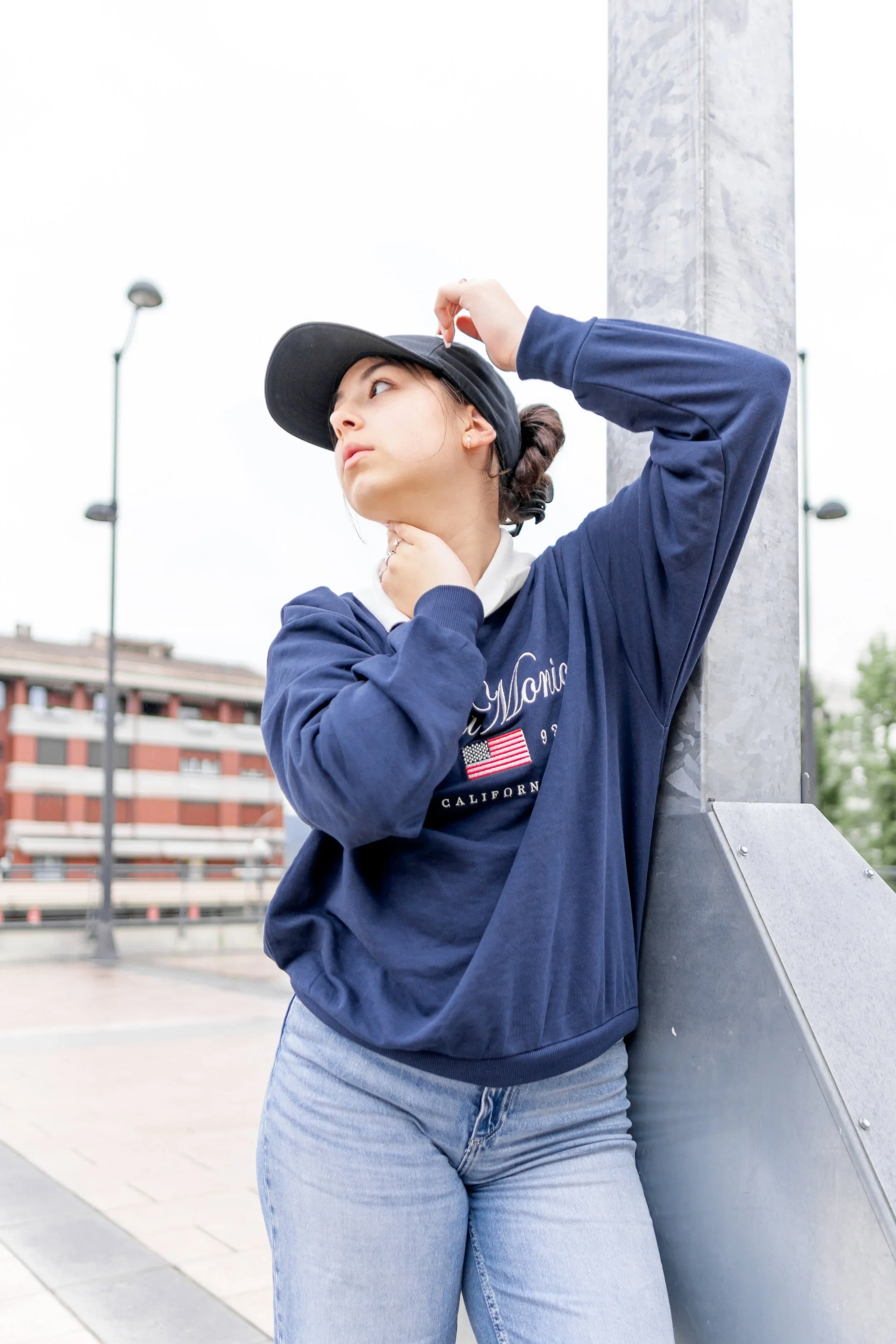 A young woman leans against a metal structure outdoors, wearing a navy blue sweatshirt with white text and an American flag, a black cap, and light blue jeans, with her hand on her neck and the other adjusting her cap.