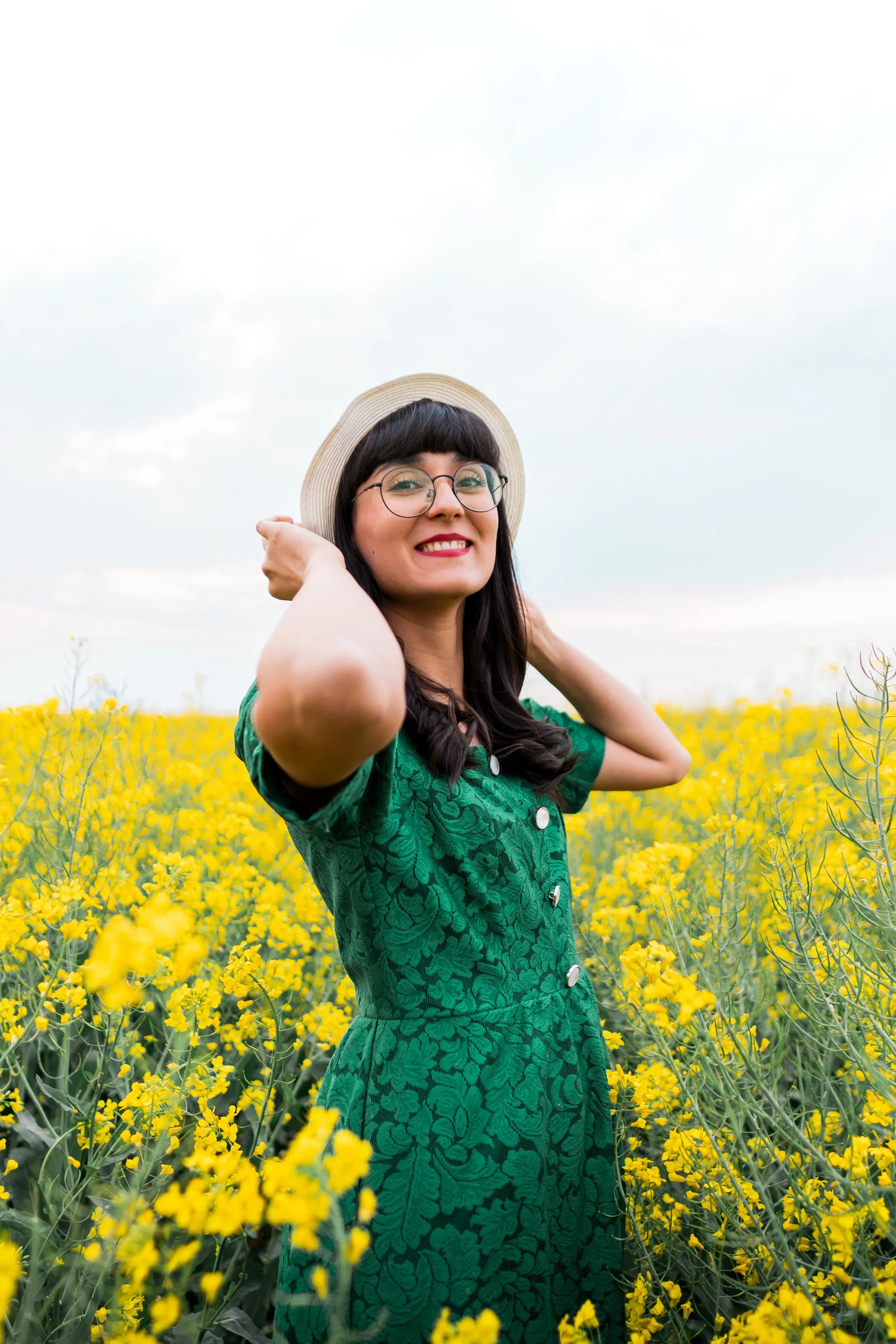 A woman in a green dress and a straw hat standing in a yellow flower field, smiling at the camera.