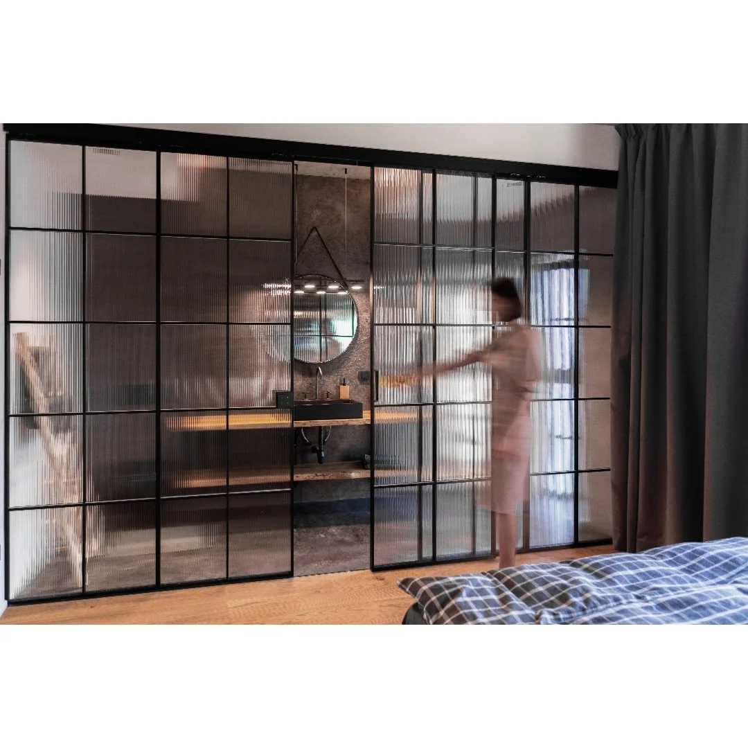 Interior of a bedroom with a glass partition wall, a person in pajamas opening the partition, a bed with a checkered blanket, a side table, a round mirror, and curtains.