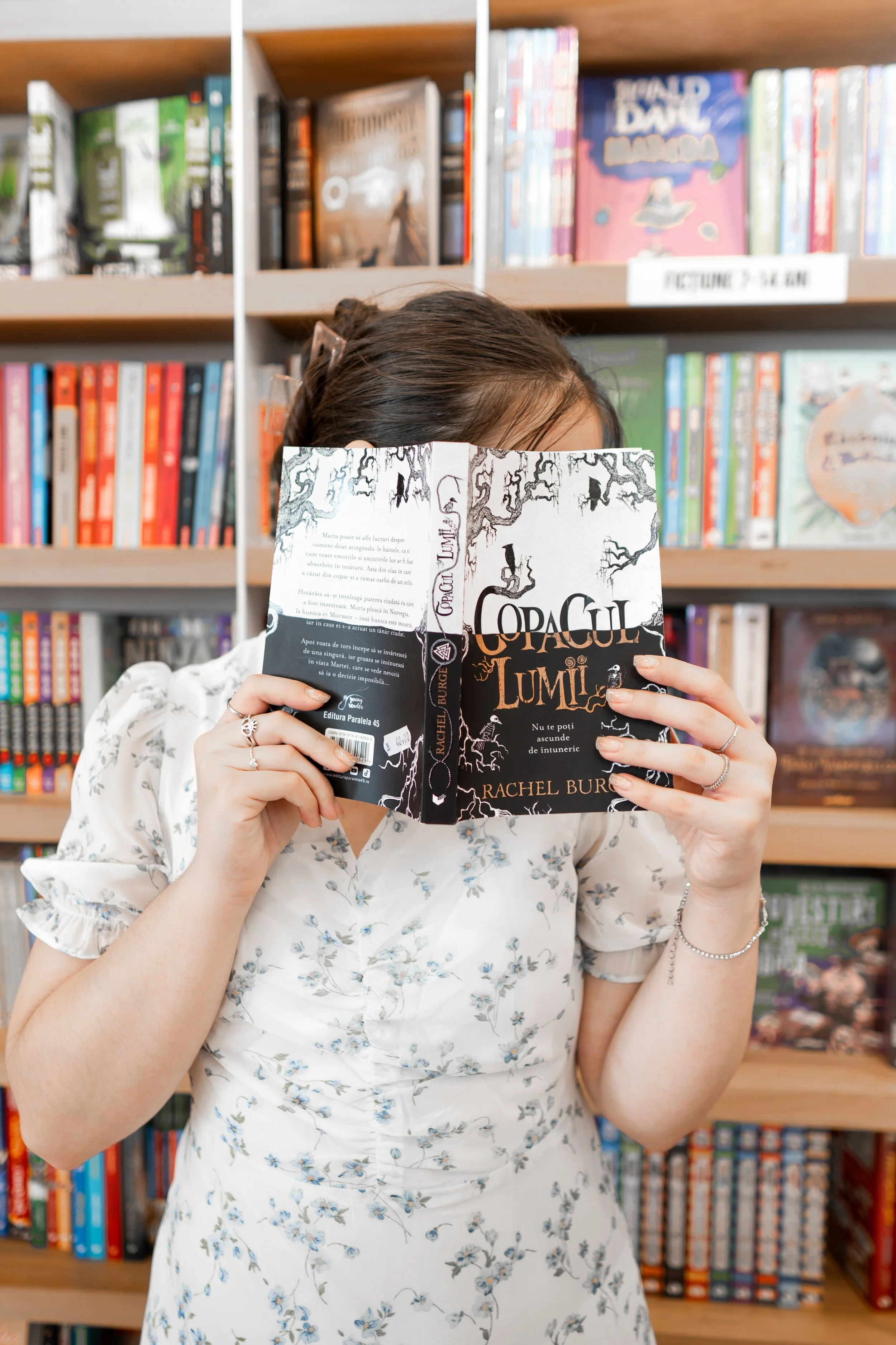 A woman holding a book titled 'Copaul Lumii' with a black and white cover featuring leafless trees and birds, in a bookstore surrounded by colorful shelves of books.