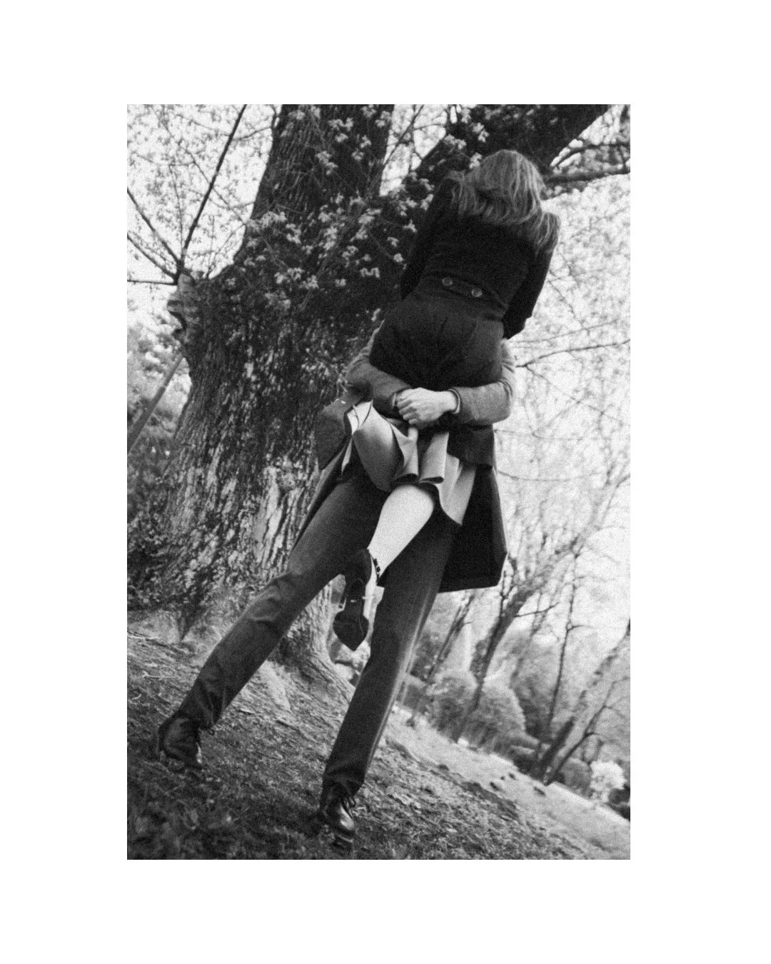 Black and white photo of two people in a park, one person standing and holding a child on their shoulders, with trees in the background.