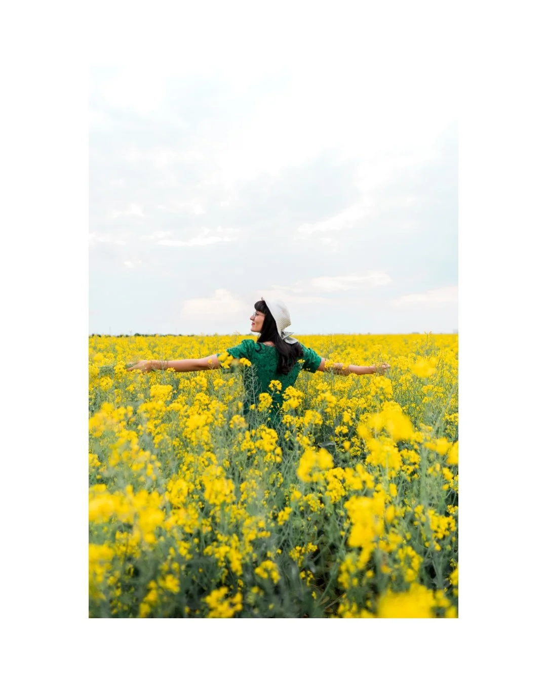 Woman in green dress with arms outstretched standing in a yellow flower field under a cloudy sky