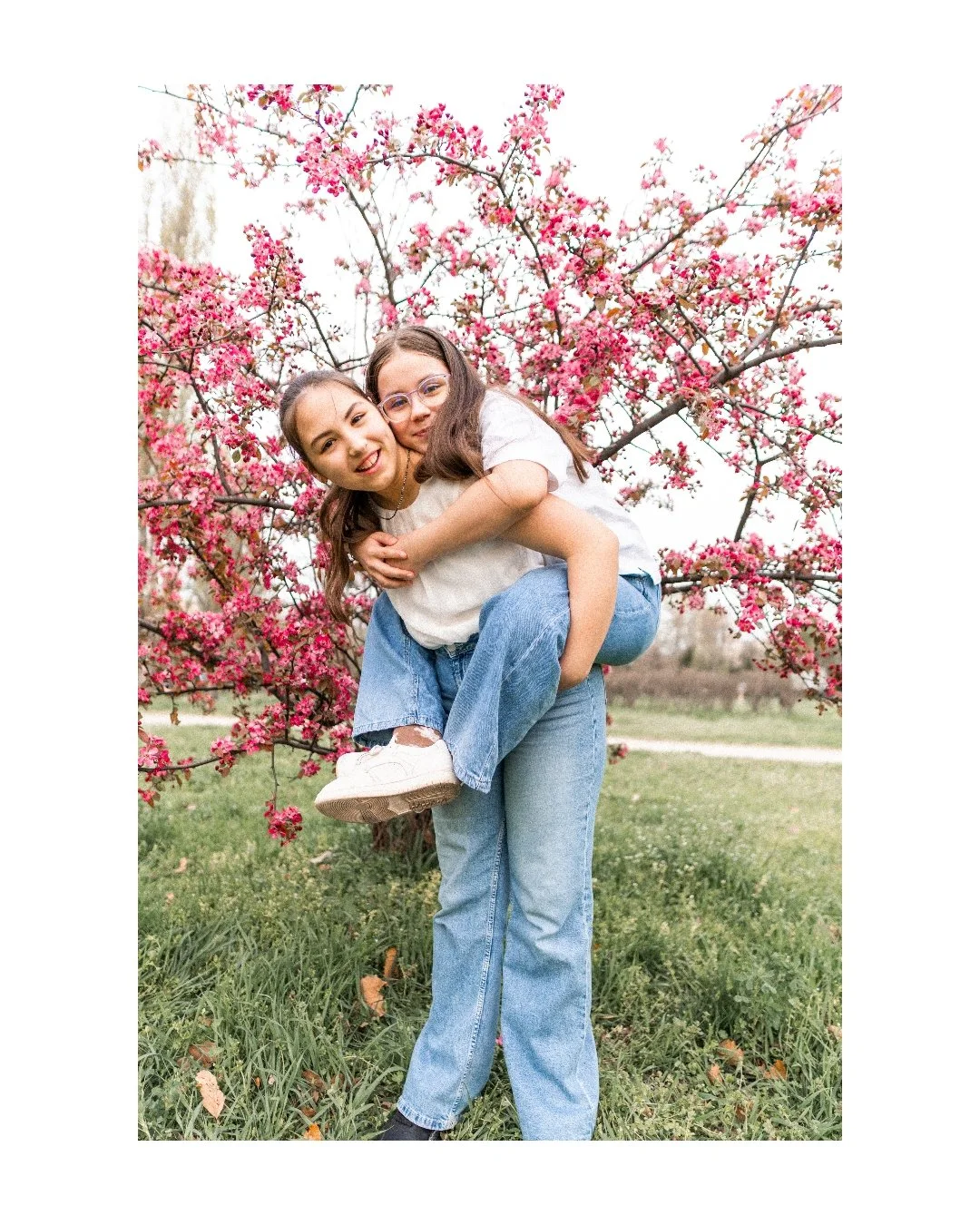 Two young girls happy and smiling, one carrying the other on her back, standing outdoors in front of a pink blossoming tree.