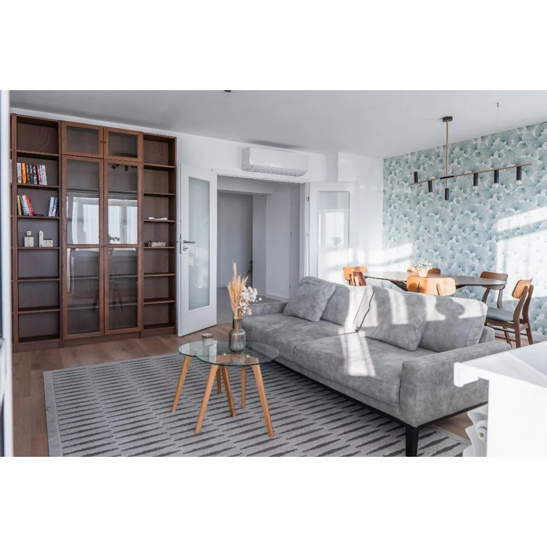 Modern living room with a gray couch, glass coffee table, wooden bookshelf, patterned wallpaper, and a dining area with wooden chairs and a flower arrangement.