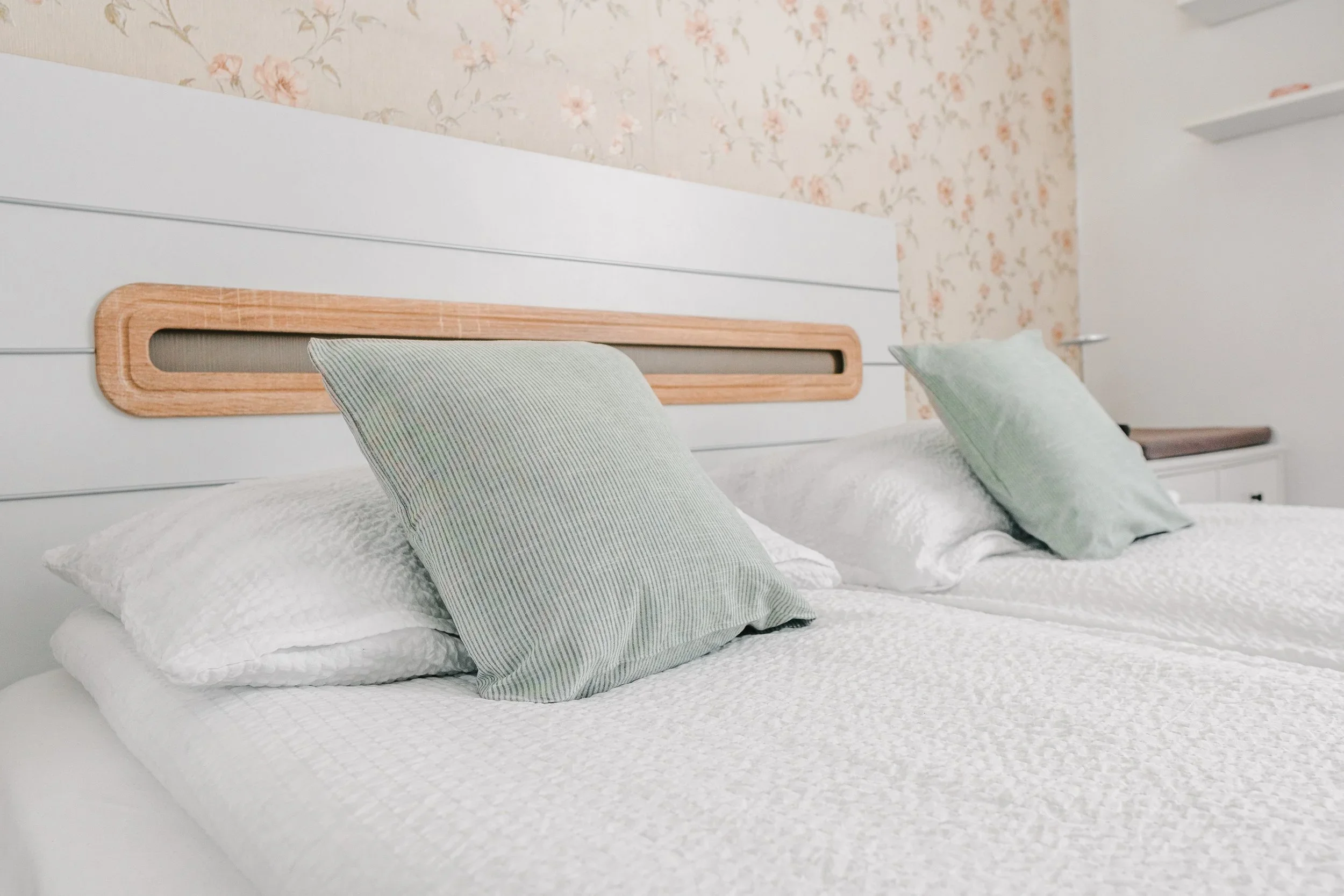 A bed with white bedding and three pillows on top, with a headboard against a floral wallpapered wall.