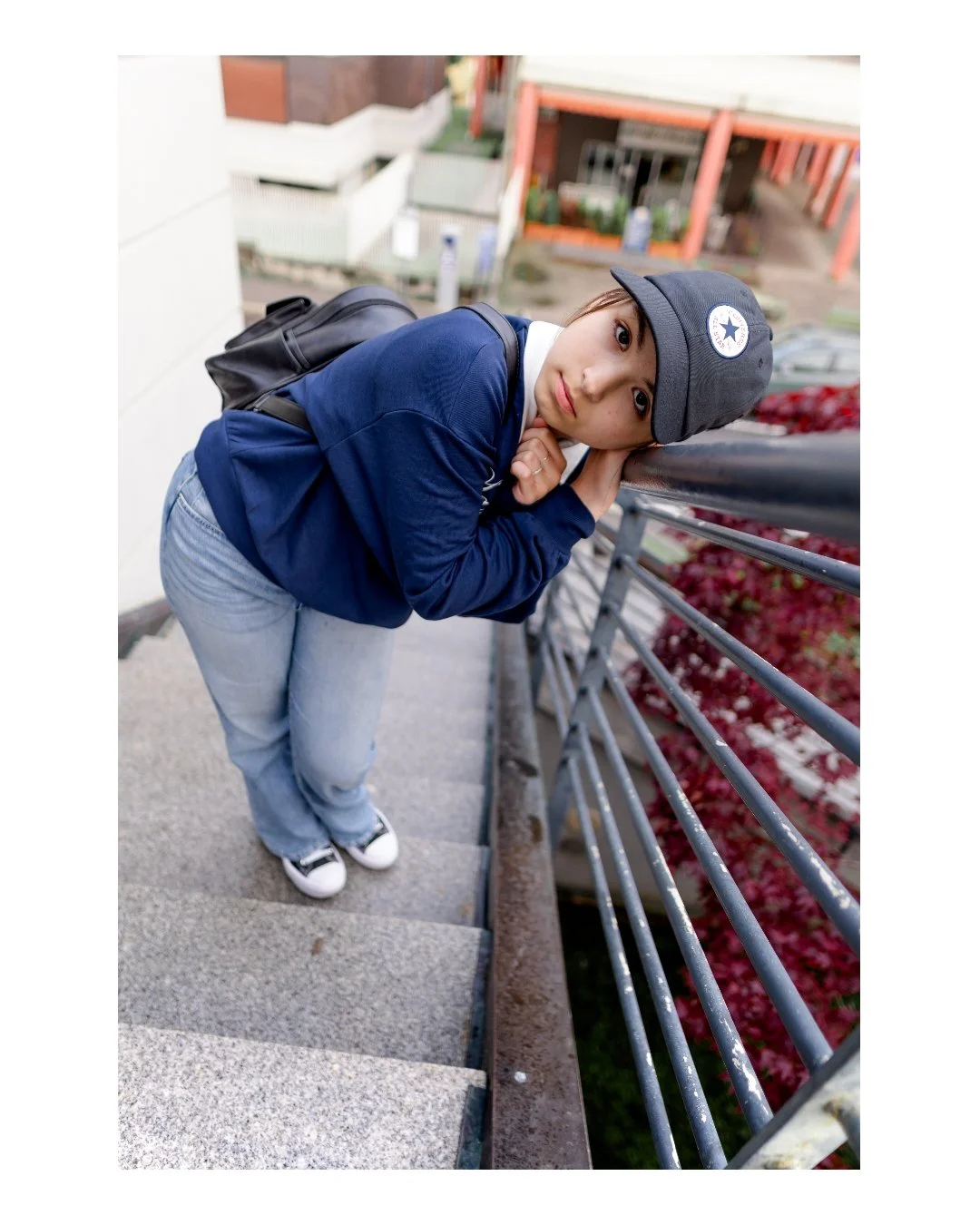 A young woman leaning over a railing on an outdoor staircase, wearing a black cap with a white logo, a blue jacket, and jeans, looking at the camera.