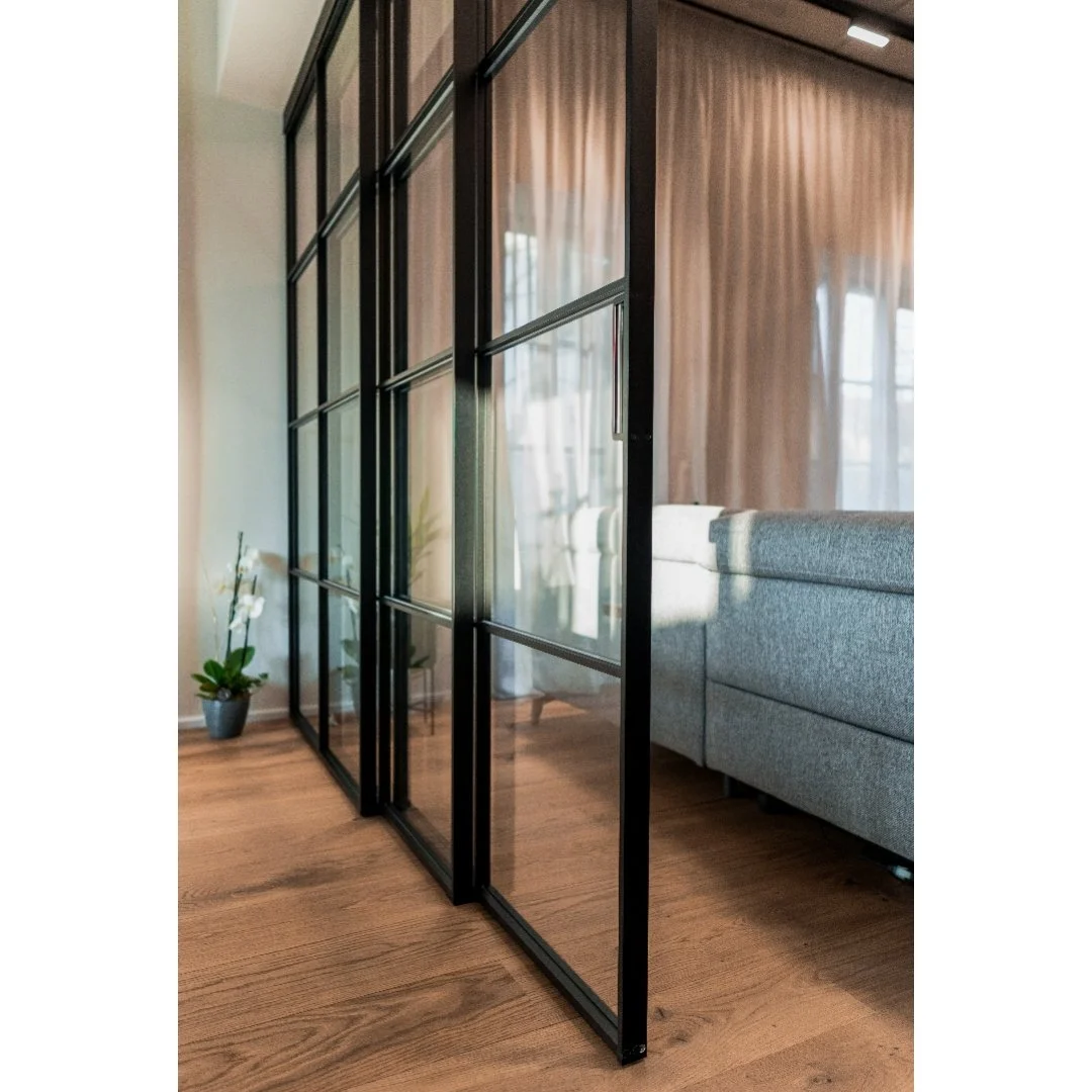 Interior view of a modern living space with a black-framed glass partition, wooden flooring, a potted orchid on the floor, and a sofa with sheer curtains in the background.