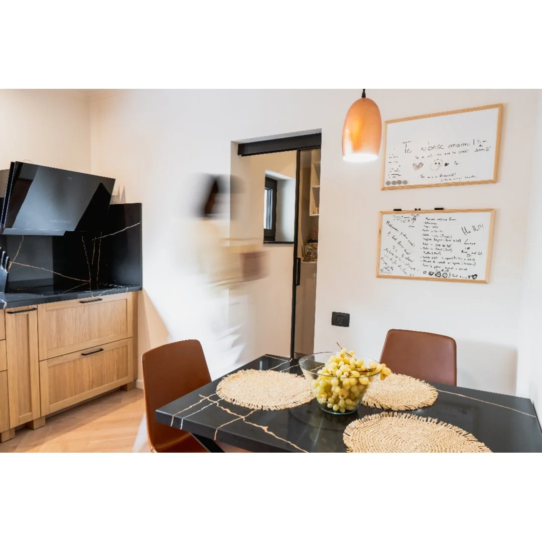 Modern dining room with a black marble table, two brown chairs, a bowl of grapes, whiteboards on the wall, a pendant light, a window, and a TV on a wooden cabinet.