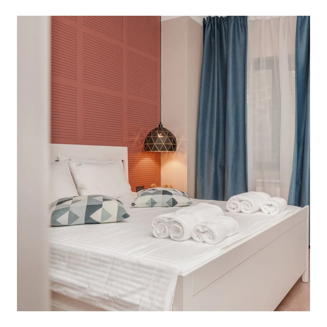 A neatly made bed with white sheets, two patterned pillows, and rolled white towels on top. The headboard is white against a salmon-colored textured wall panel. To the right, there are blue curtains partially covering a window, and a black geometric pendant lamp hangs beside the bed.