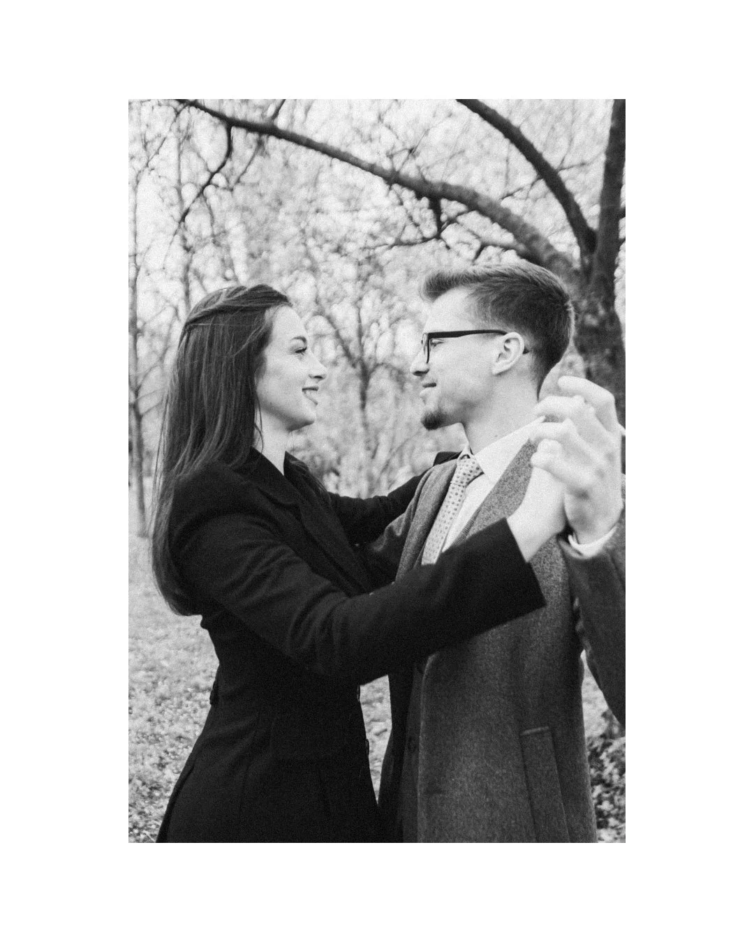A black and white photo of a couple dancing outdoors, facing each other and smiling, with trees in the background.
