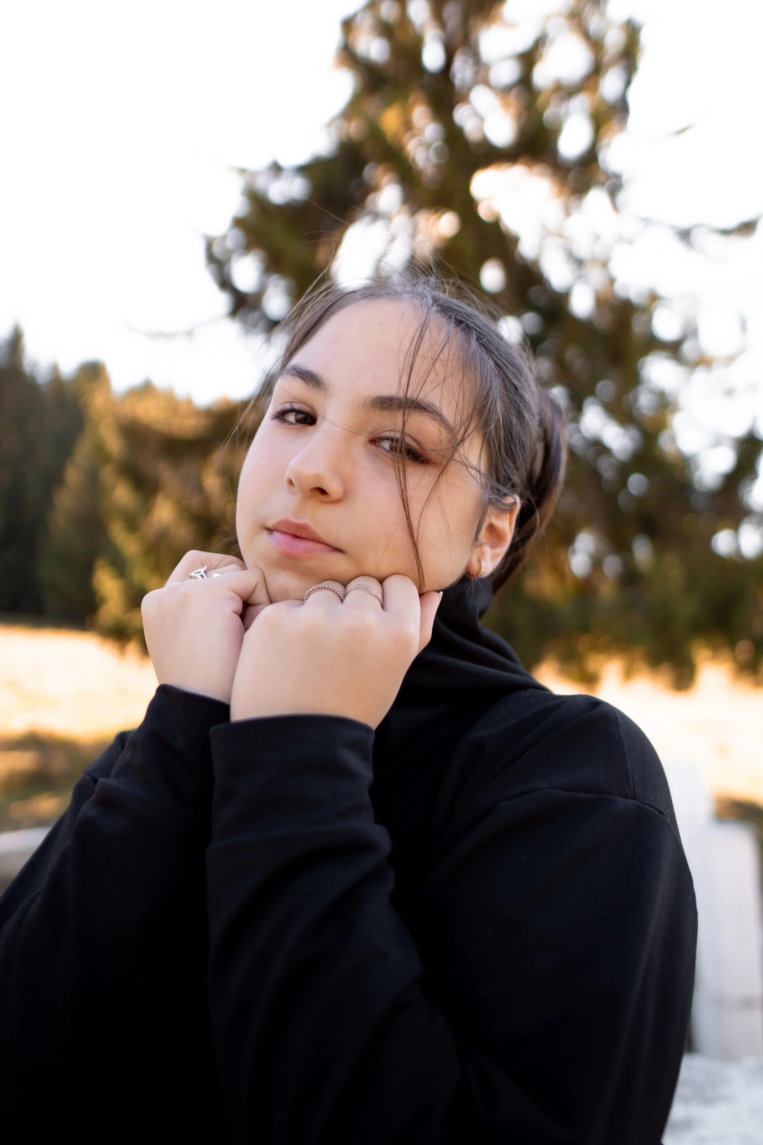 A young woman with dark hair, wearing a black hoodie, resting her chin on her hands, outdoors in a natural setting with blurred trees in the background.