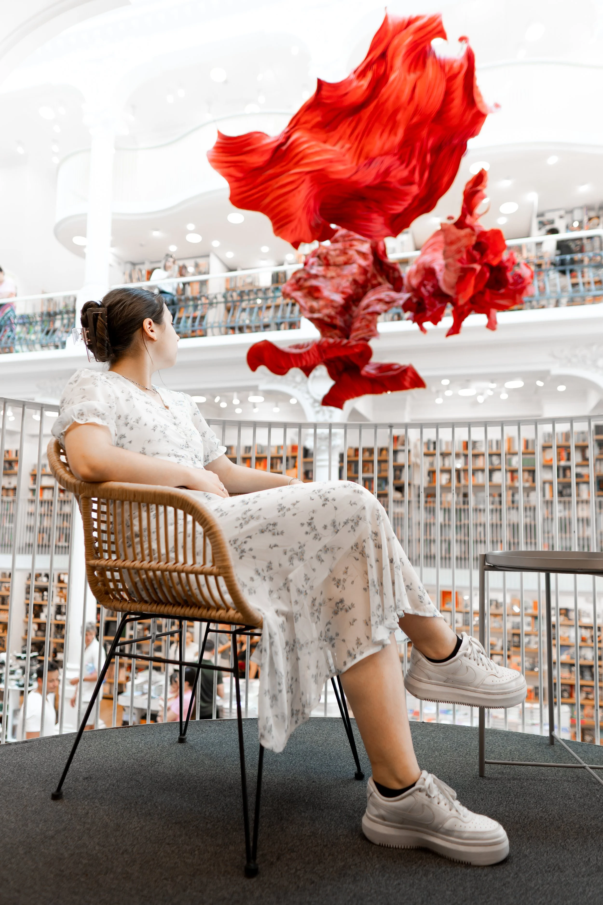 A woman in a white floral dress and white sneakers sitting on a chair at an indoor library or bookstore, observing large red abstract floral art hanging from the ceiling.