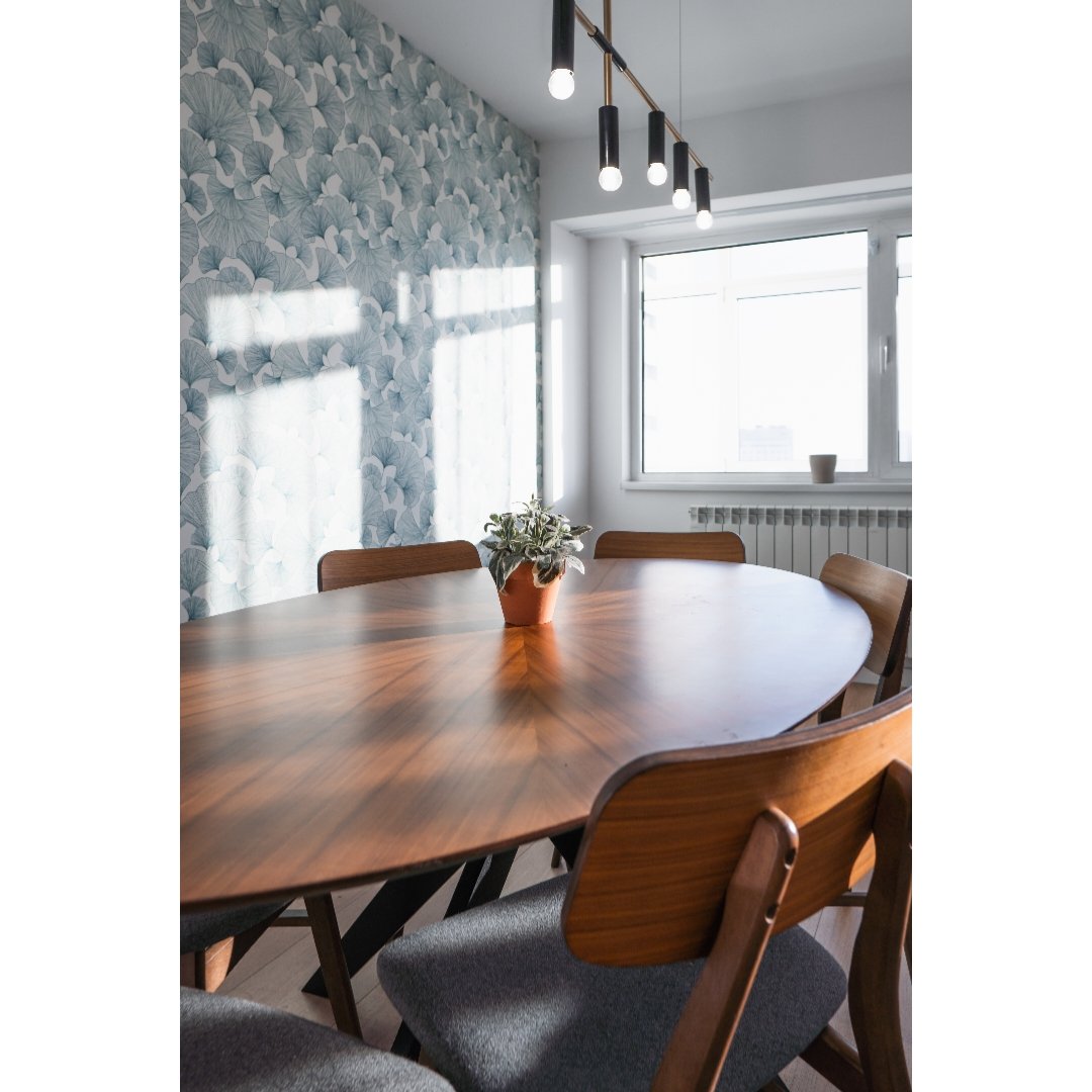 Dining room with a wooden oval table, six wooden chairs with gray cushions, a potted plant in the center, a window with sunlight, floral wallpaper on one wall, and hanging light fixtures.