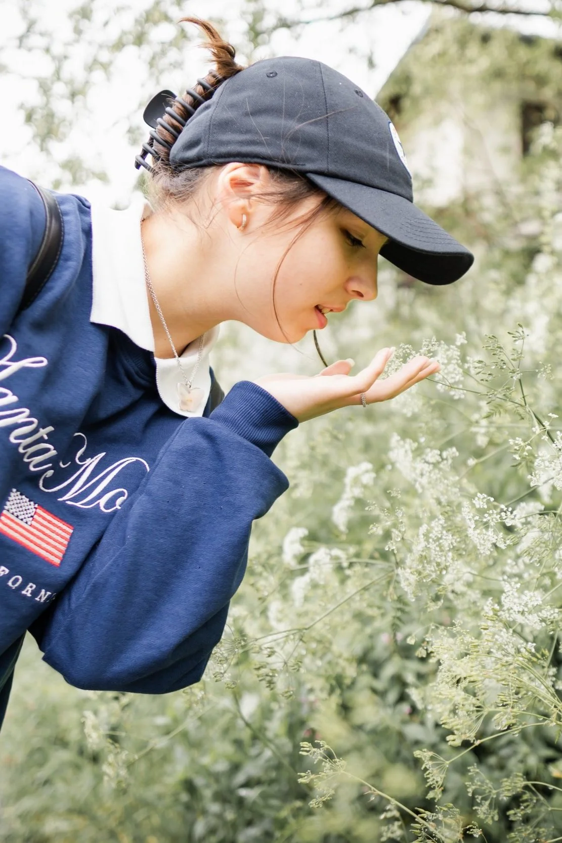 A young woman wearing a black cap, a blue sweatshirt, and a white collar, bending down outdoors to smell or examine small white flowers on a plant.