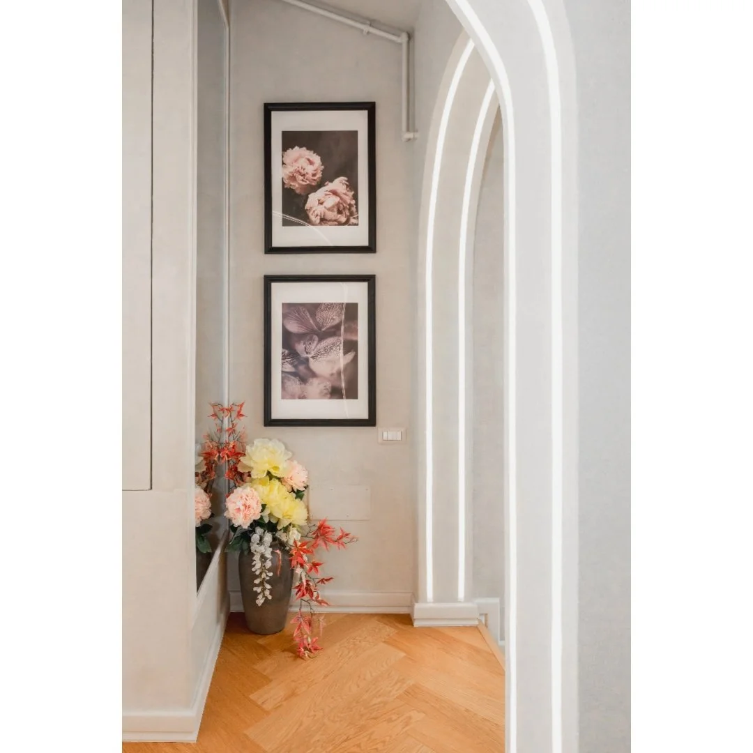 Corner of a room with a large floral arrangement in a vase, two framed floral photographs on the wall, and an arched doorway.