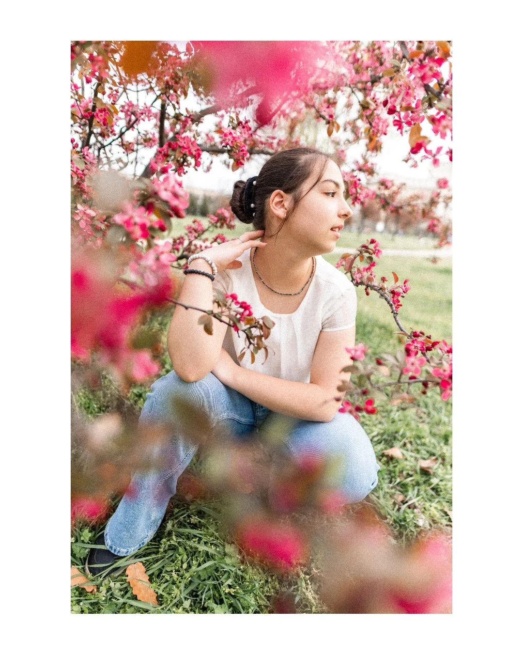 A young woman with dark hair tied in a bun, wearing a white t-shirt and jewelry, sitting among pink flowering trees outdoors.
