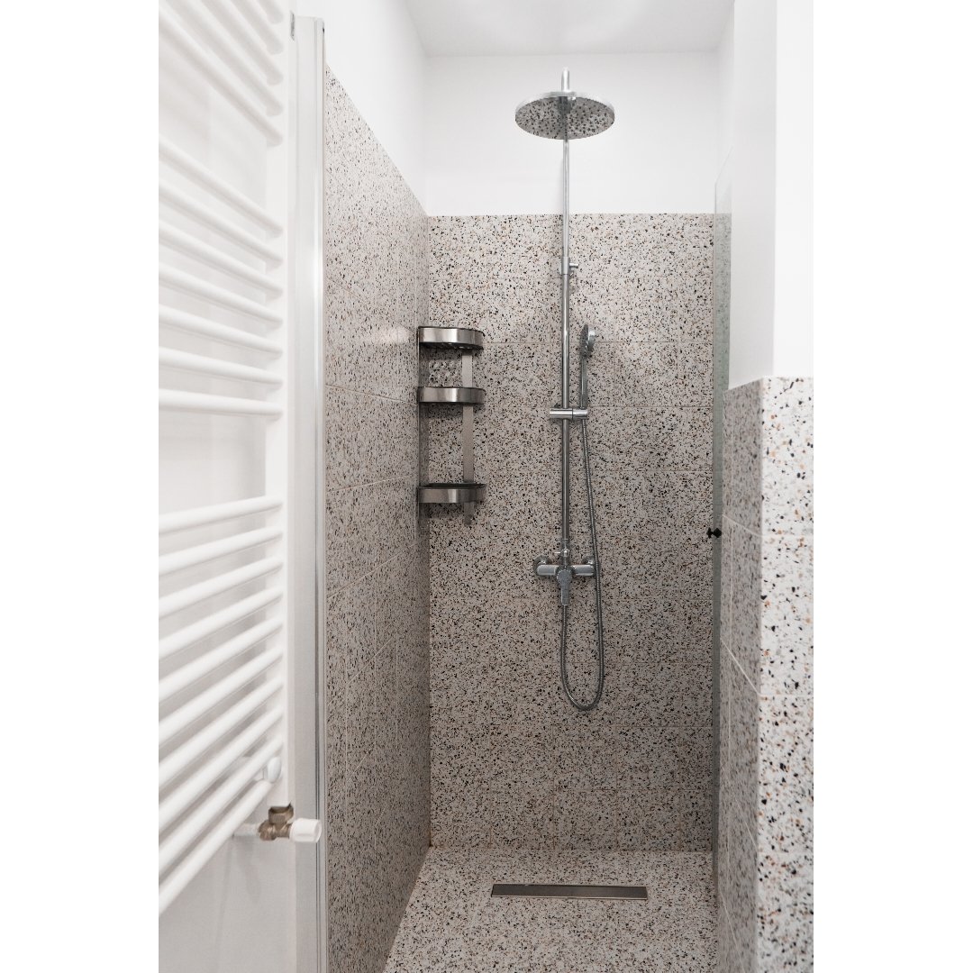 Shower with a circular overhead showerhead, a handheld shower fixture, and a three-tier metal corner shelf, with speckled tile walls and floor.