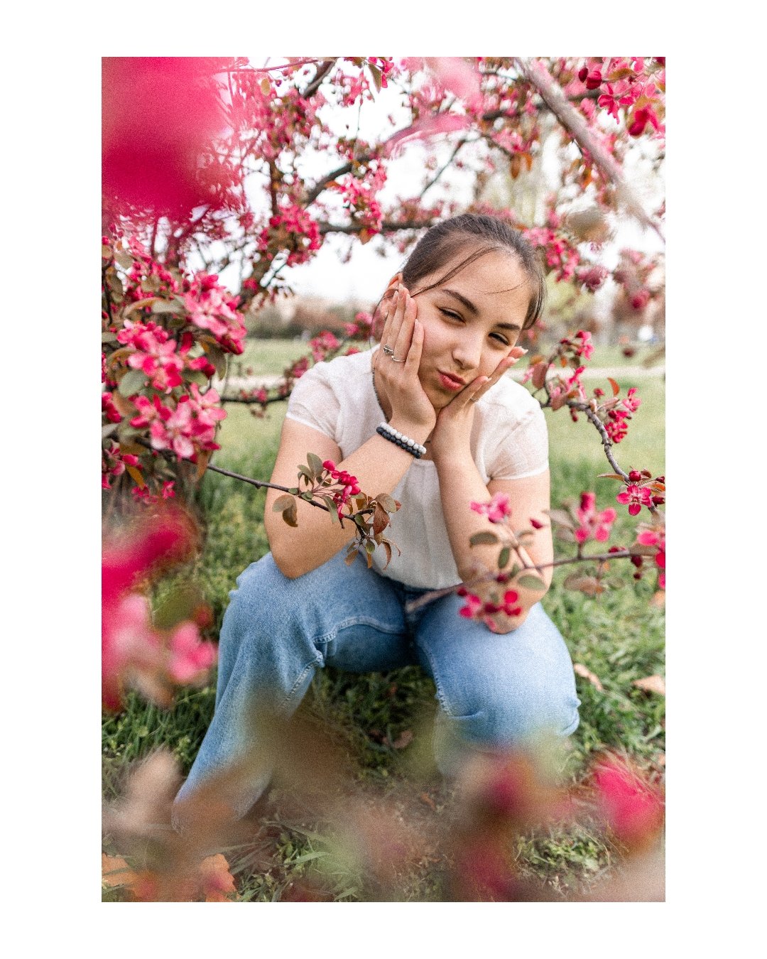 Young woman squatting among blooming pink flowers on tree branches, holding her face with both hands and making a playful expression.