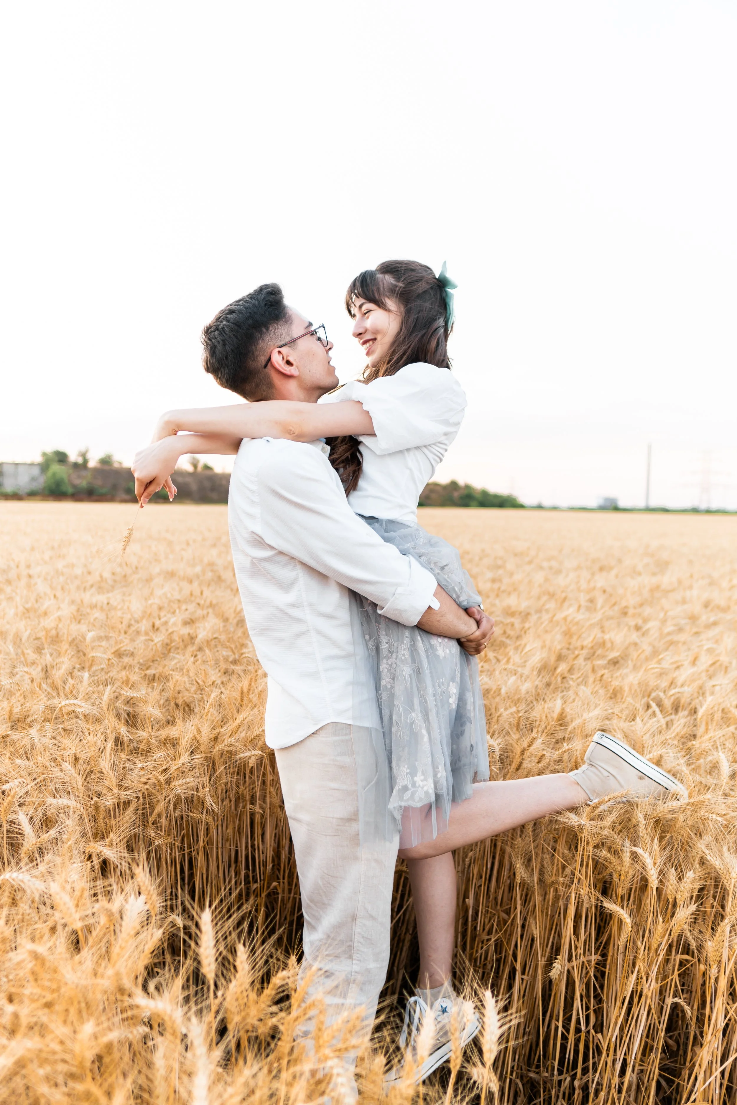A man holding a woman in a wheat field, both smiling and gazing at each other, with the woman's arm around his neck and one leg lifted, during daytime.
