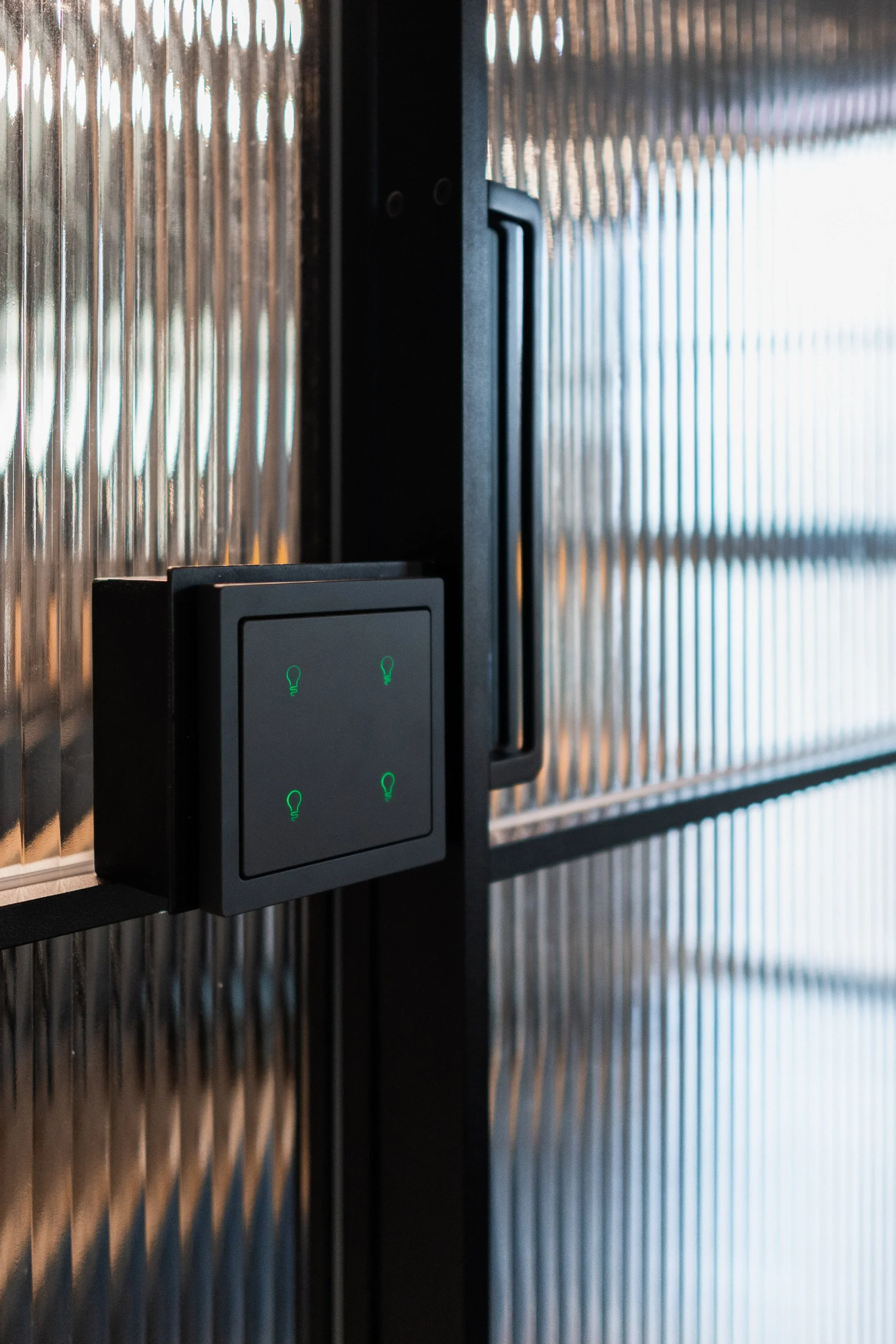 Close-up of a black control panel with four green illuminated light bulb icons, mounted on a textured metallic wall.
