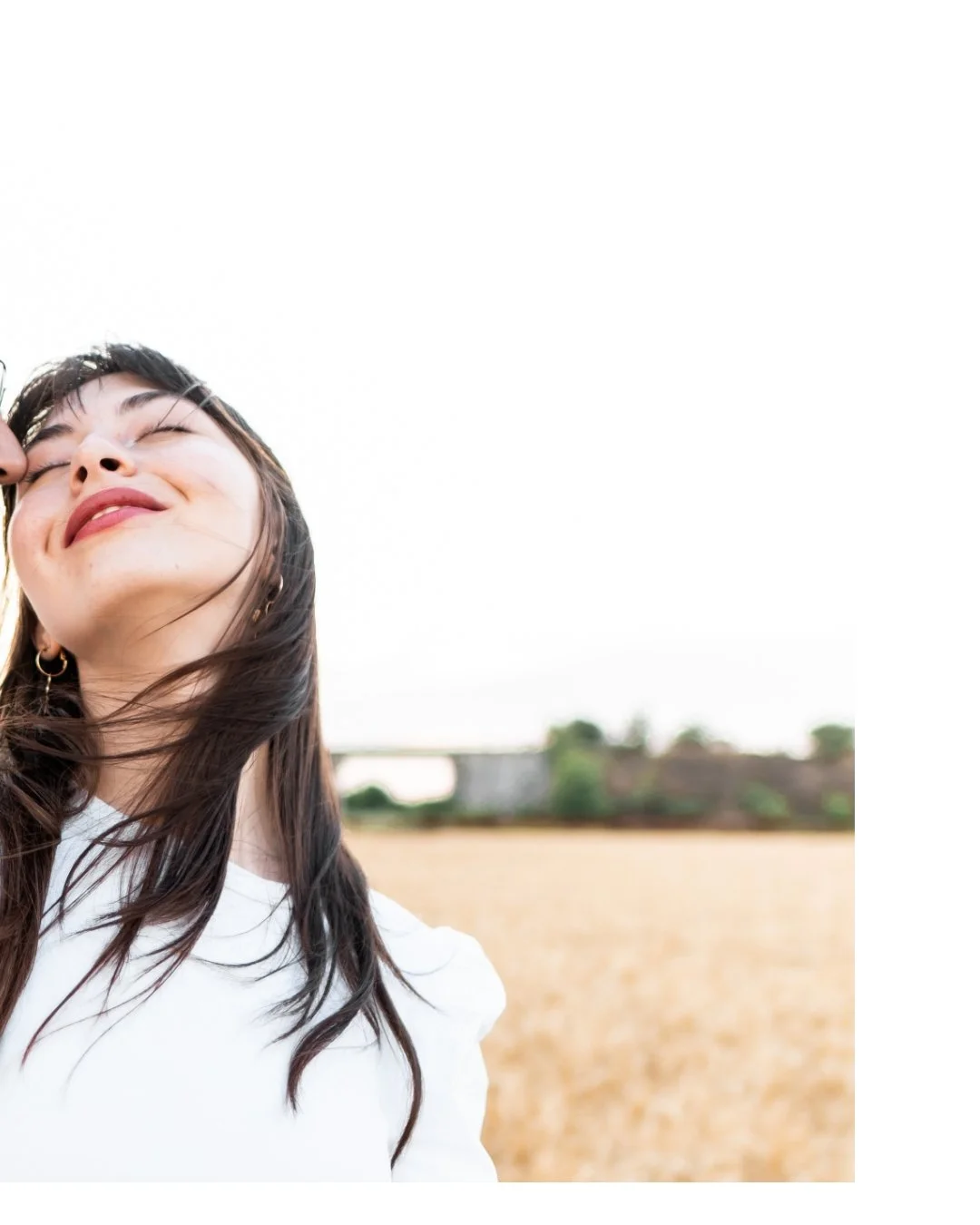 A woman with long dark hair smiling with her eyes closed outdoors in a field with trees and a bridge in the background.