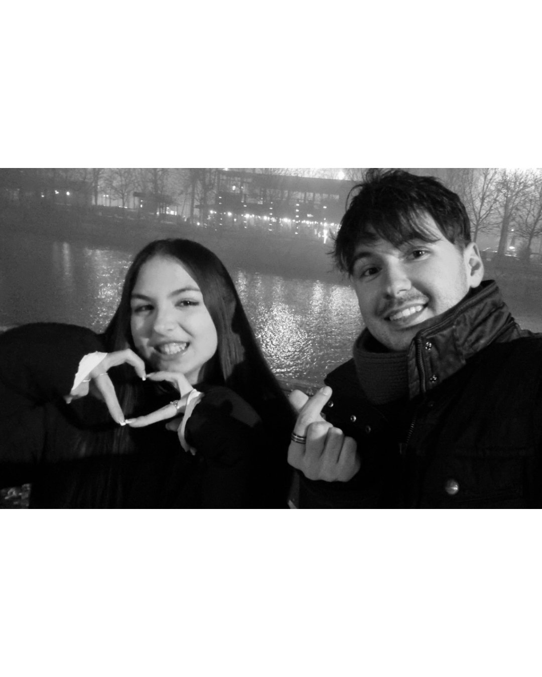 Black and white photo of a young couple smiling outdoors near water, with trees and park in background. The woman makes a heart shape with her hands, and the man is pointing at her.