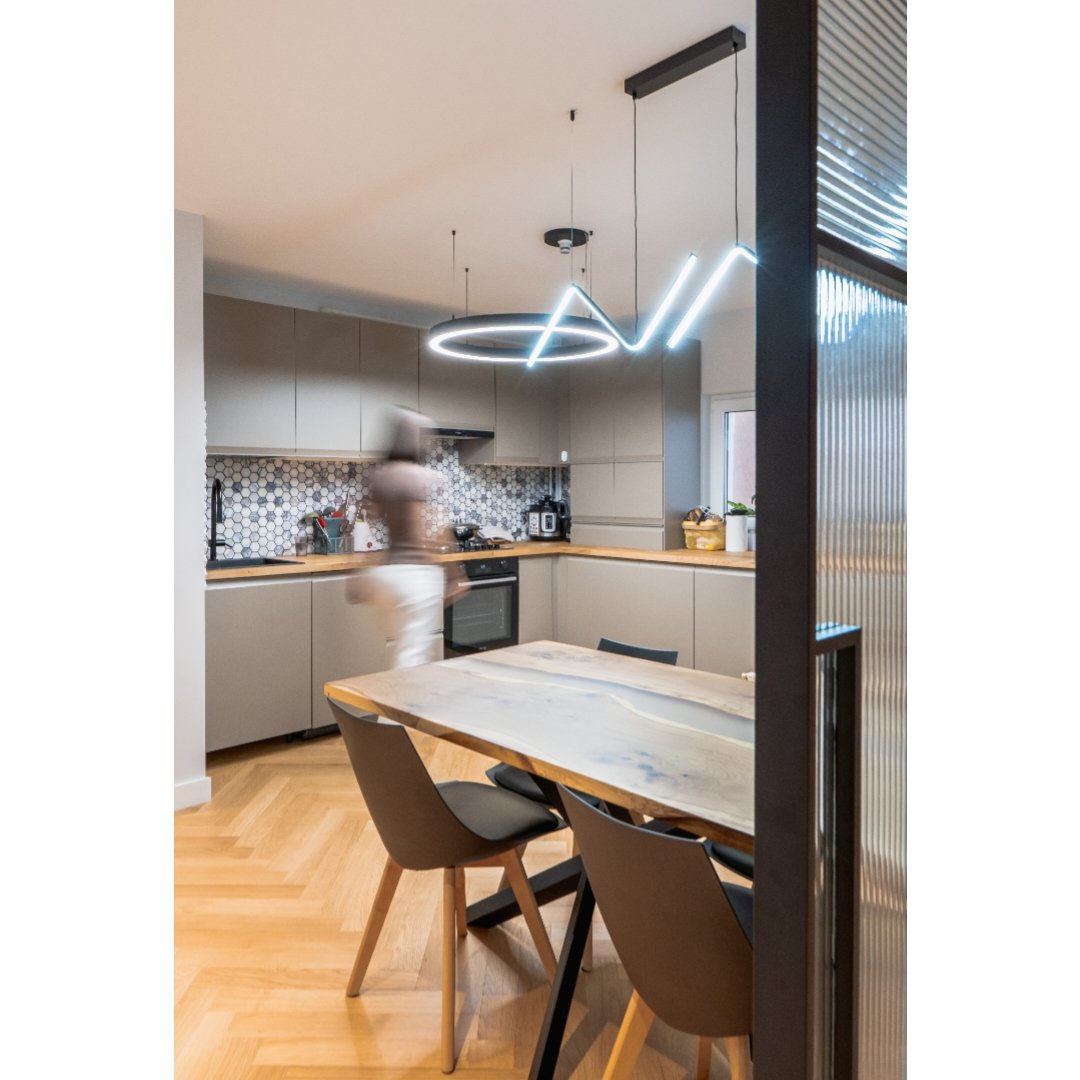 Modern kitchen with beige cabinets, hexagon tile backsplash, and a wooden dining table with black chairs. Hanging abstract LED light fixture includes circular and diagonal elements. A person is blurred near the countertop.
