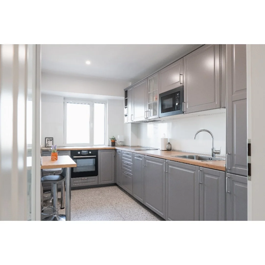 Modern kitchen with gray cabinets, a built-in microwave, a black oven, a wooden countertop, a sink with a faucet, and a small breakfast table with two stools near a window.