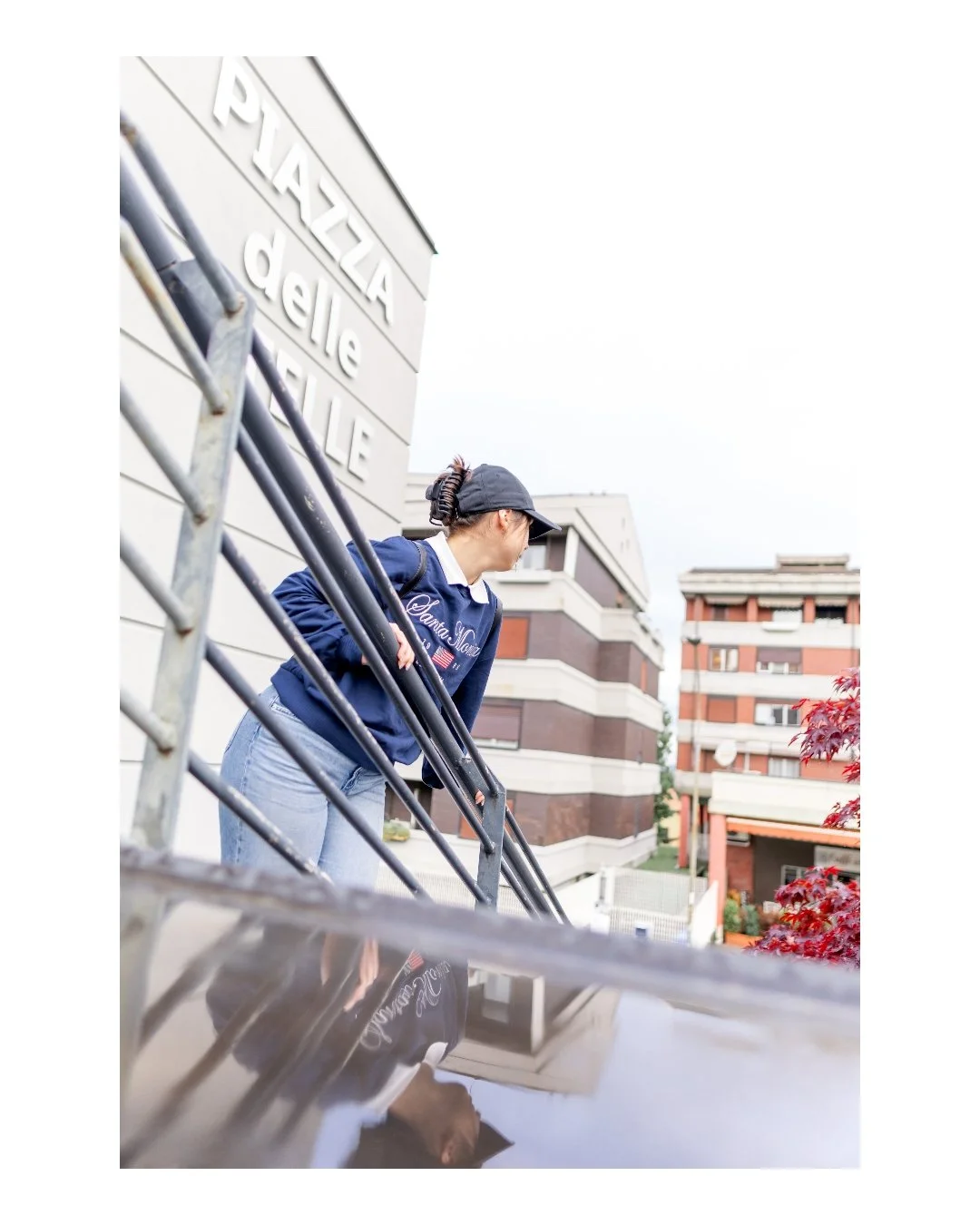 A woman wearing a baseball cap, sunglasses, and a navy sweatshirt with text, standing outside near a building with a sign that reads 'PIZZA' and 'deli,' leaning on a metal railing, and looking to the right.