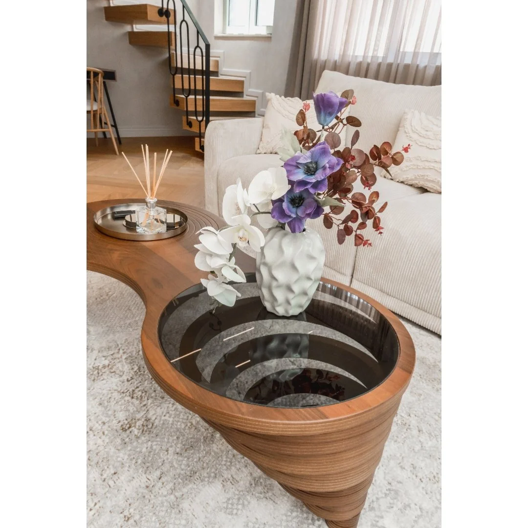 A living room with a wooden coffee table featuring a round black glass inset, holding a white textured vase with purple and white artificial flowers. Behind the table, there is a cream-colored sofa with decorative white pillows, a beige curtain covering a window, and a staircase with wooden steps and black iron balusters.