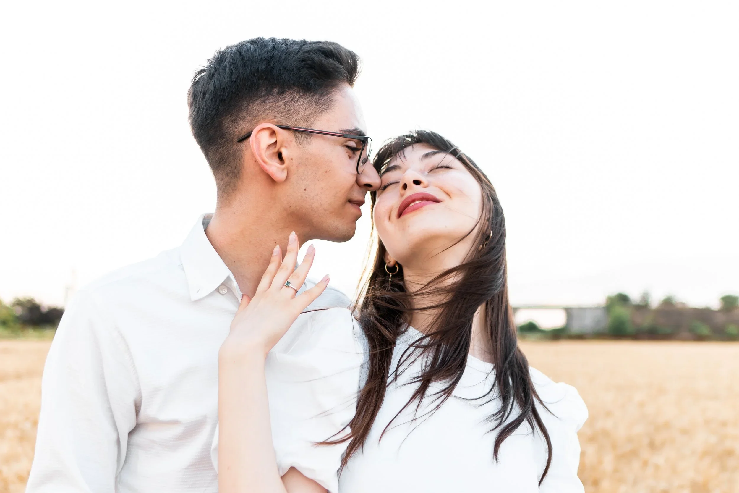 A young couple standing close together outdoors, with the man gently touching the woman's neck and both smiling with eyes closed, in a field with a bridge in the background.