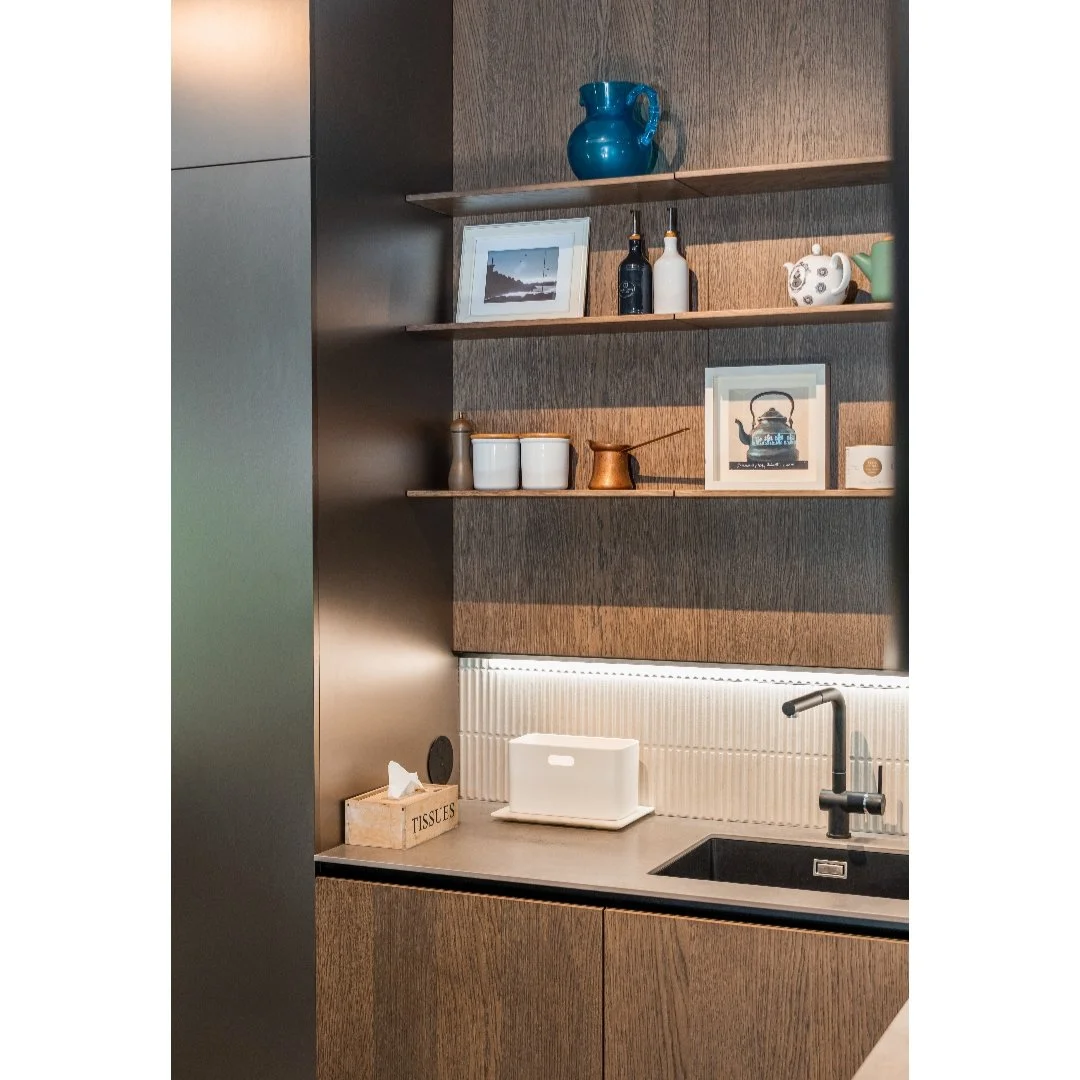 Kitchen with wooden and metallic cabinets, open shelves holding framed photos, vases, and teapots, each shelf displaying decorative items, with a sink, paper tissue box, and tissue packet on the countertop.
