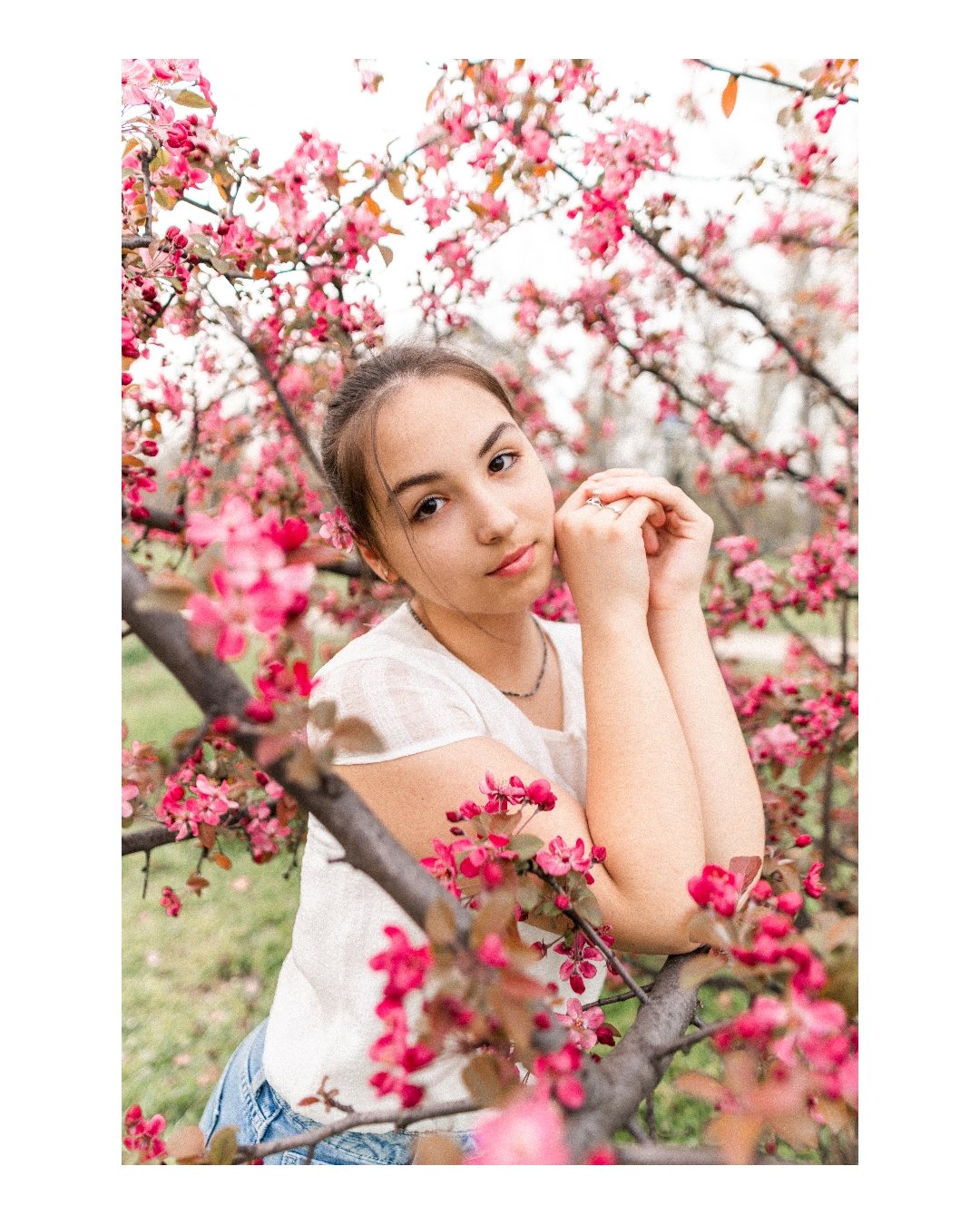 A young woman with brown hair, wearing a white top, posing among pink flowering branches outdoors.