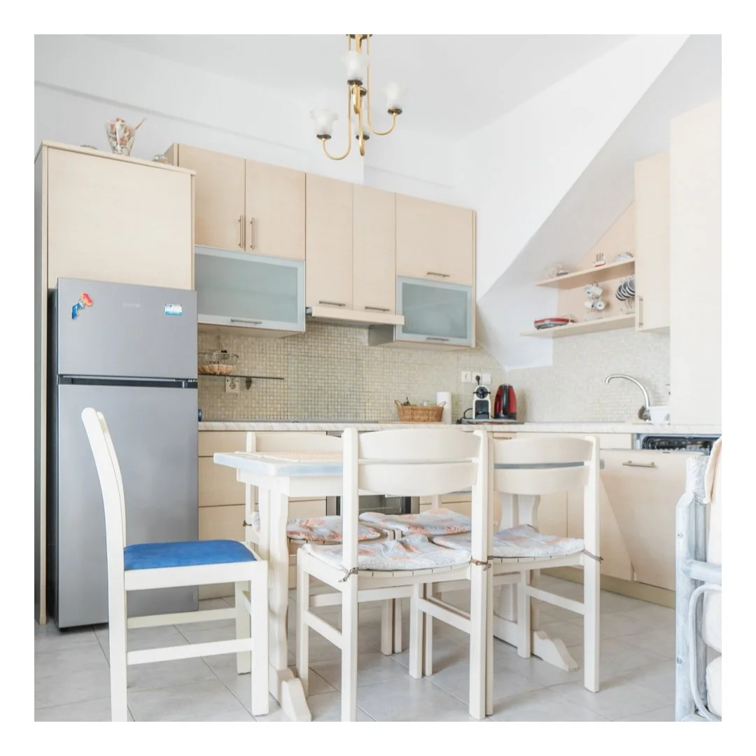 A kitchen with beige cabinets, stainless steel refrigerator, white dining table with four chairs, and a chandelier overhead. Includes appliances on the countertop and open shelves with dishes.