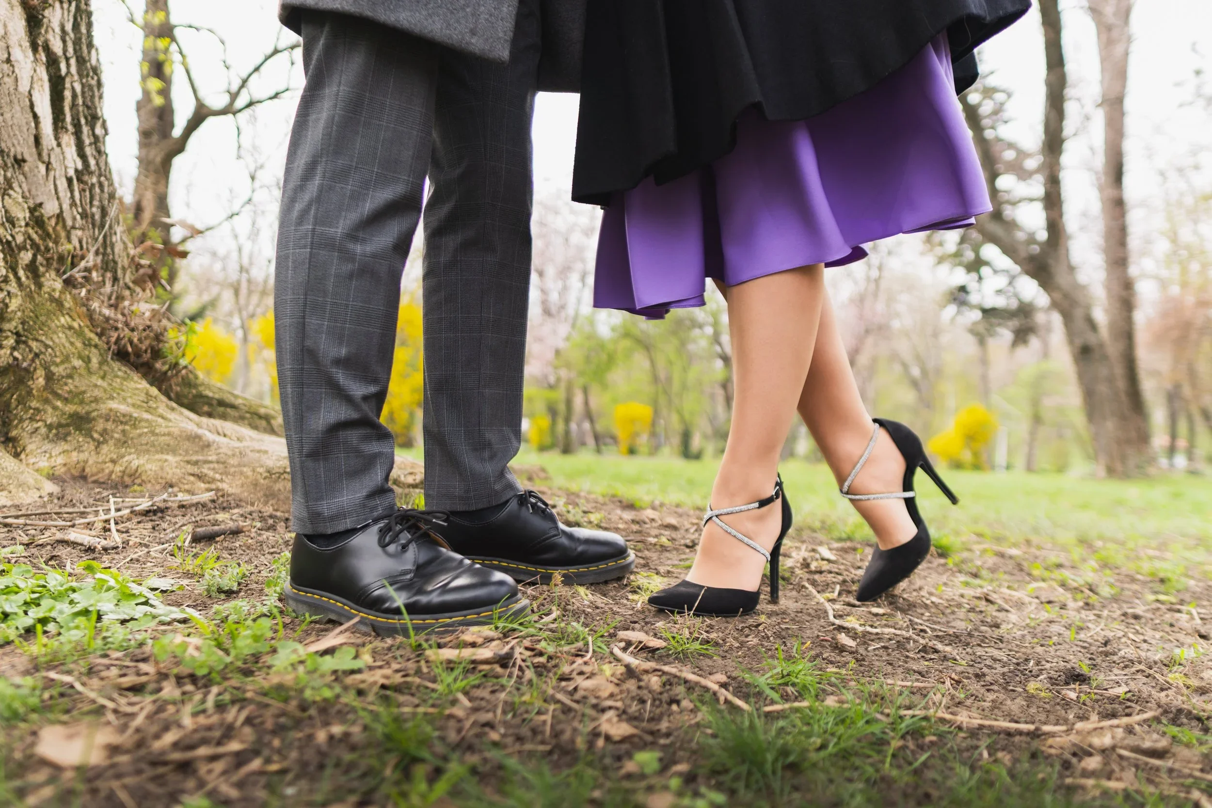 Close-up of a man wearing black shoes and gray plaid pants and a woman wearing black high heels with ankle straps and a purple skirt standing outdoors on grass and dirt near a tree.