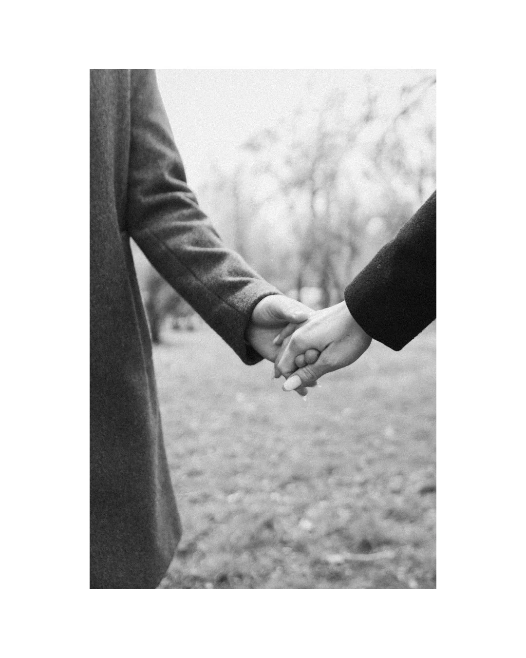 Two people holding hands outdoors, one in a long coat and the other in a darker coat, in black-and-white.