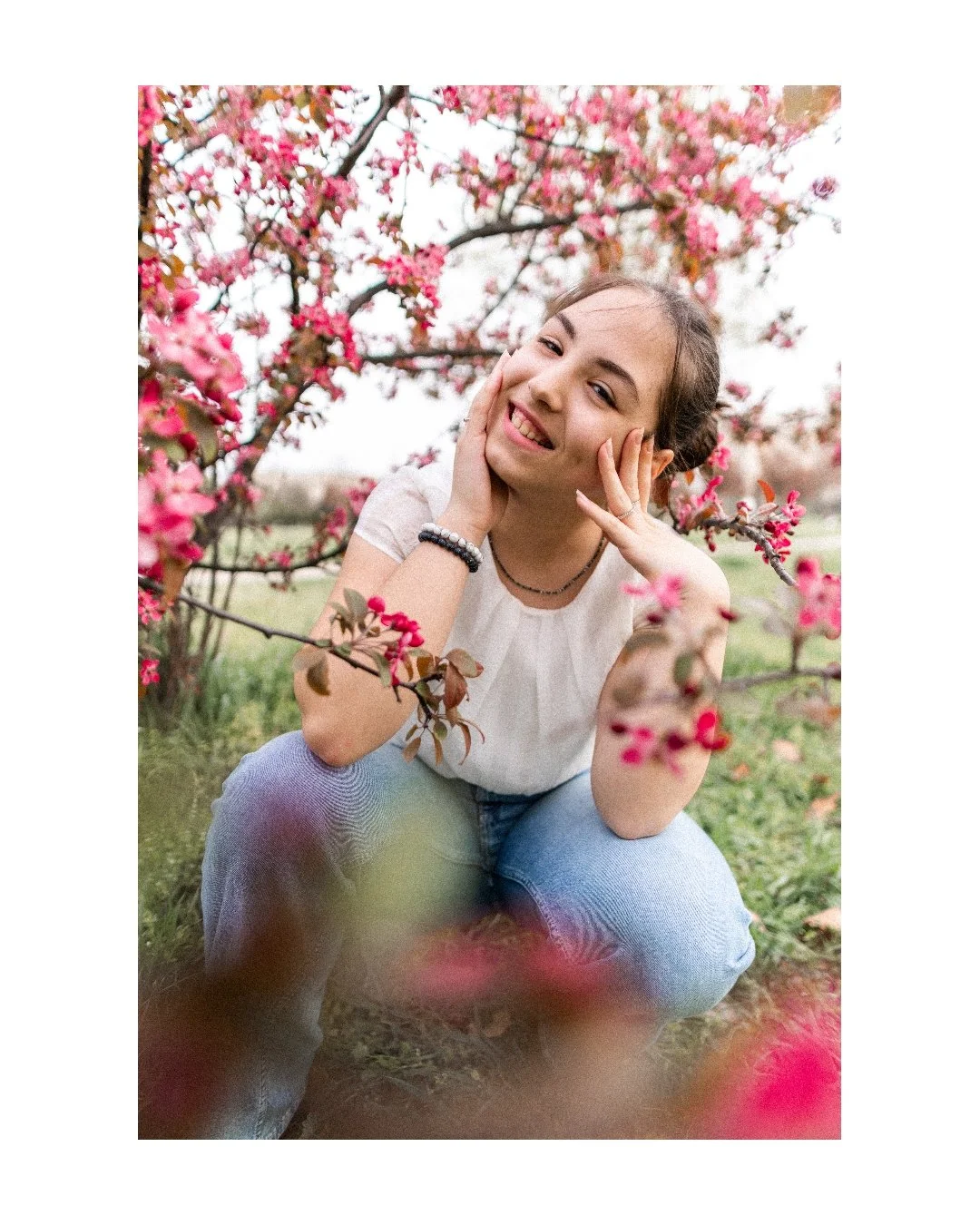 A young woman with dark hair, smiling and squatting among pink flowering branches, wearing a white t-shirt, jeans, and bracelets.