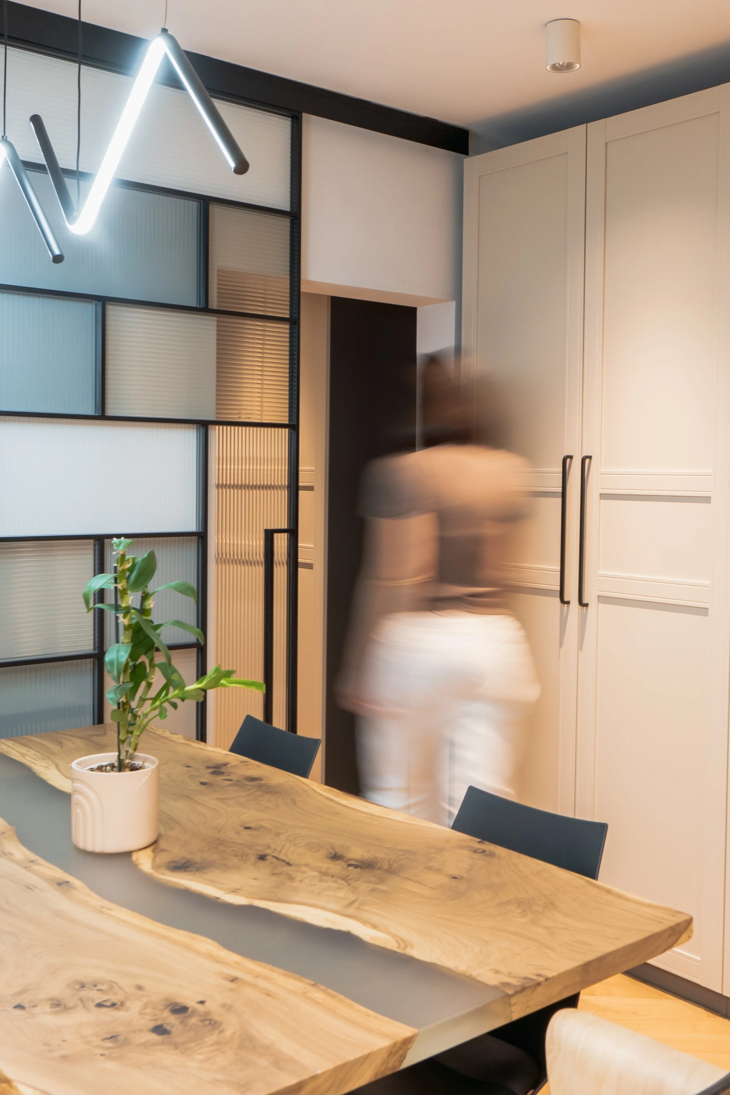 A blurred figure of a person standing in a modern dining room with a wooden table, black chairs, a potted plant, a glass partition, and white cabinets, with contemporary lighting fixtures.